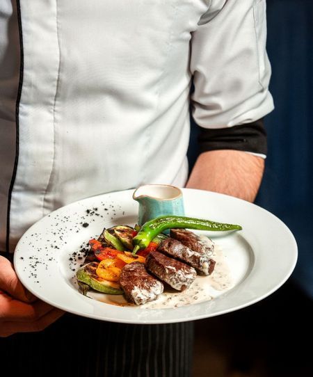 Chef holding a plate with grilled meat, vegetables, sauce, and pepper.