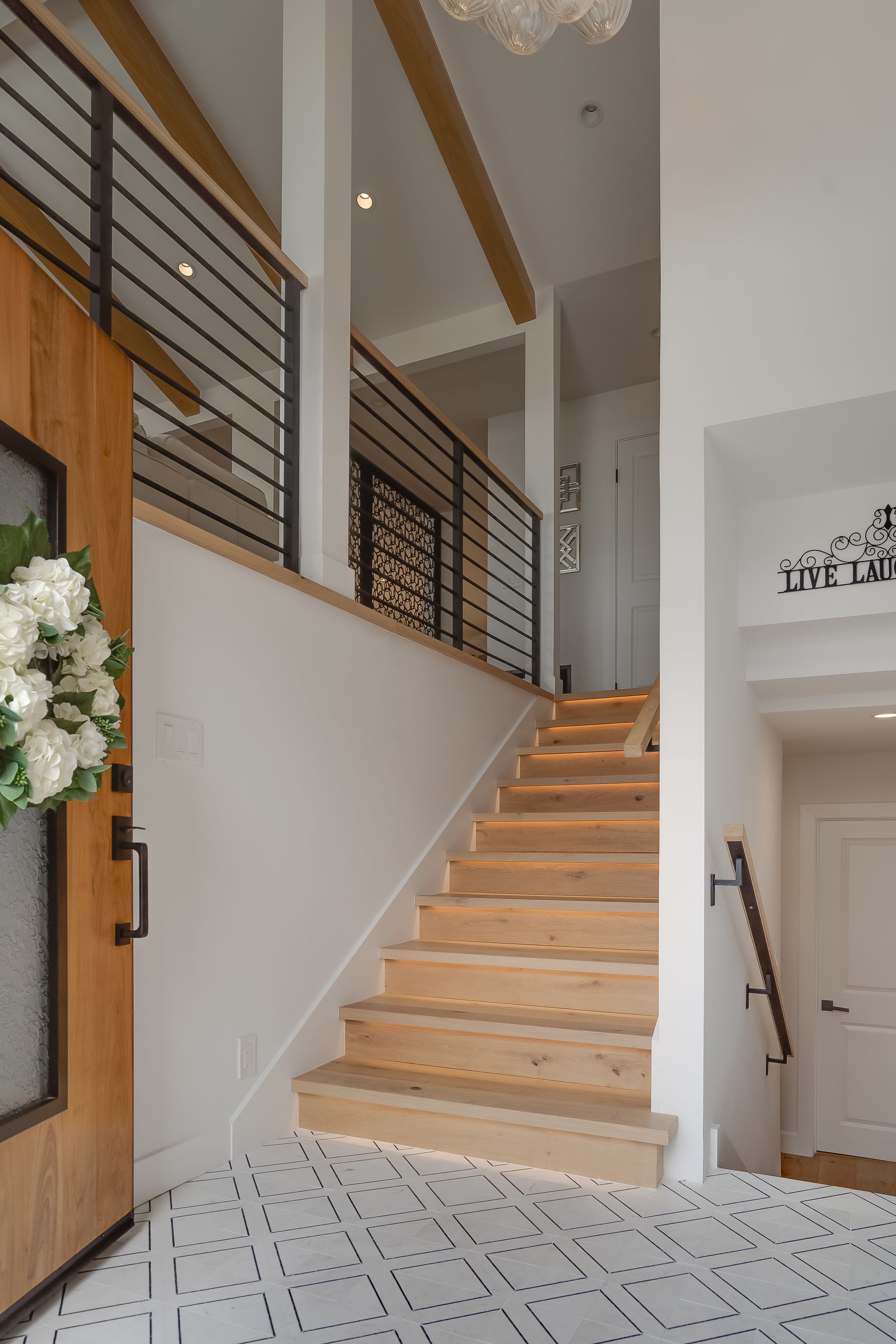 Wooden staircase in a modern home with a light-filled entry, black railing, and patterned tile floor.