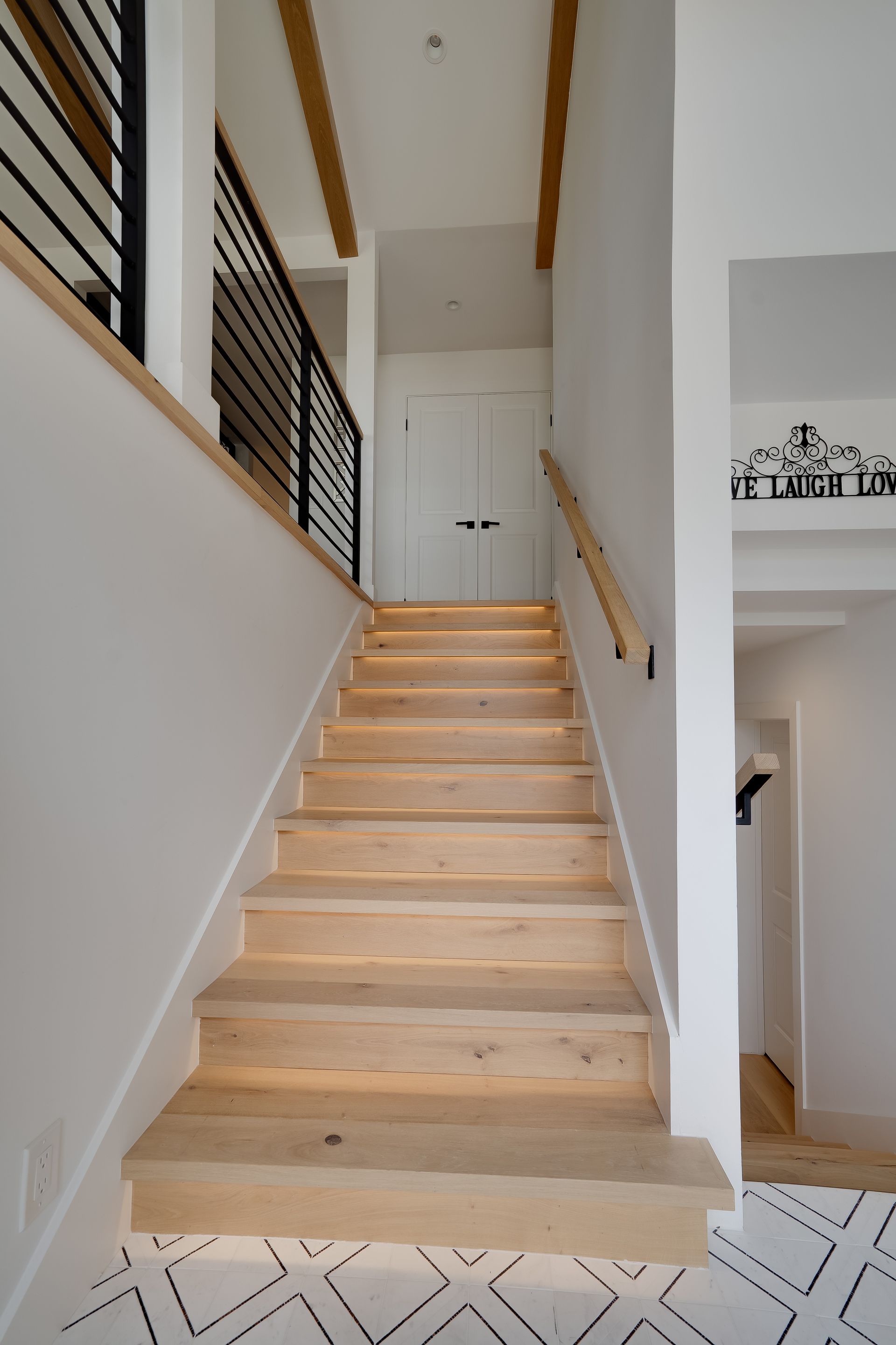 Wooden staircase with light fixtures, white walls, black metal railing, and a doorway.