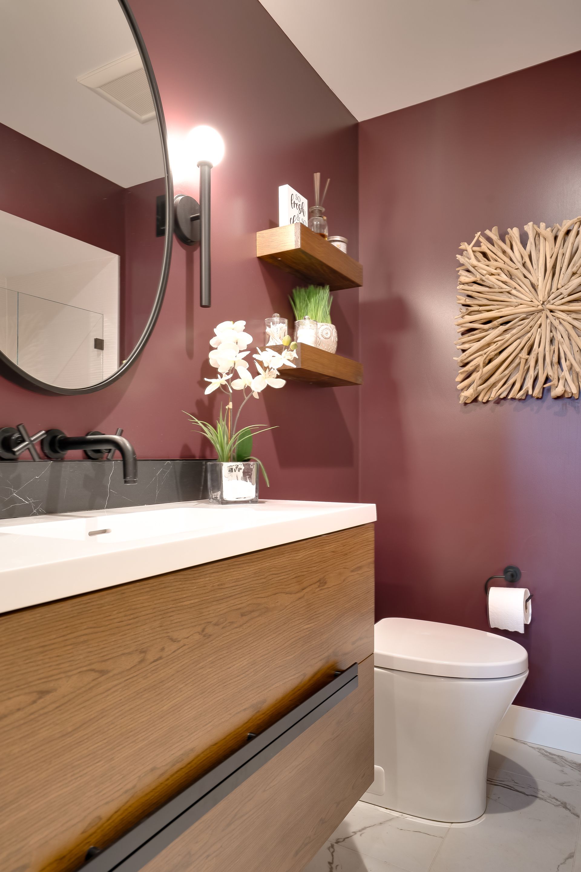 Bathroom with burgundy walls, wooden vanity and floating shelves, round mirror, and toilet.