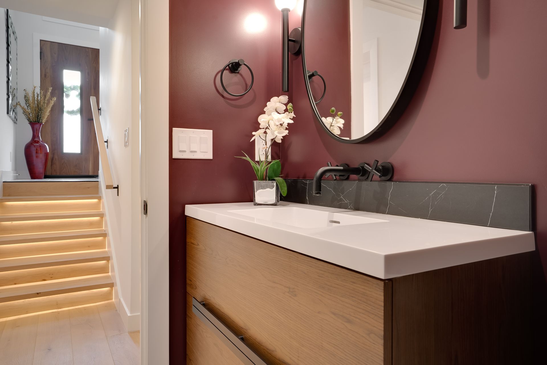 Modern bathroom with burgundy wall, wooden vanity, round mirror, and stairs leading to a door.