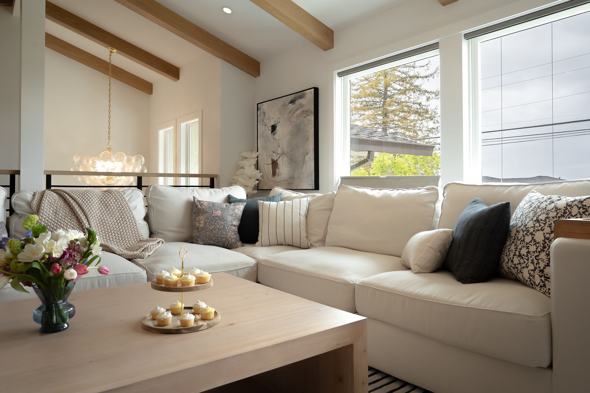 Cozy living room with white sectional sofa, wooden beams, and a coffee table with treats and flowers.