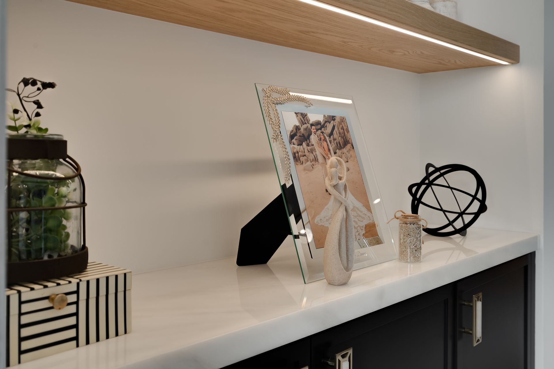 Shelf with framed photo, jar, and black decorative items. White wall and black cabinet with wood shelf.