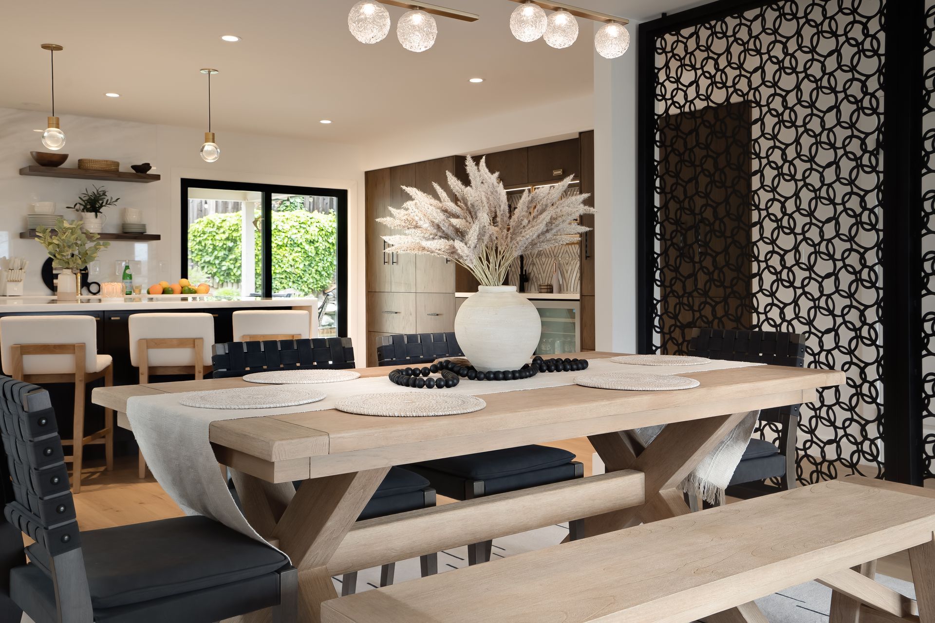 Dining room: light wood table with bench and chairs, white vase with pampas grass, open to kitchen with bar seating.