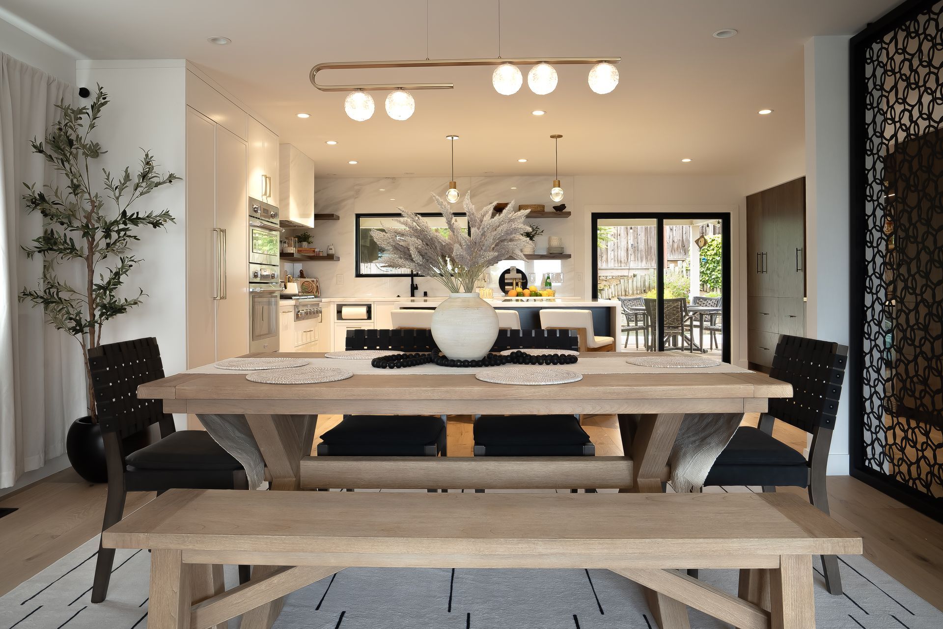 Dining room with a long wooden table, bench, and black chairs. Kitchen in the background.