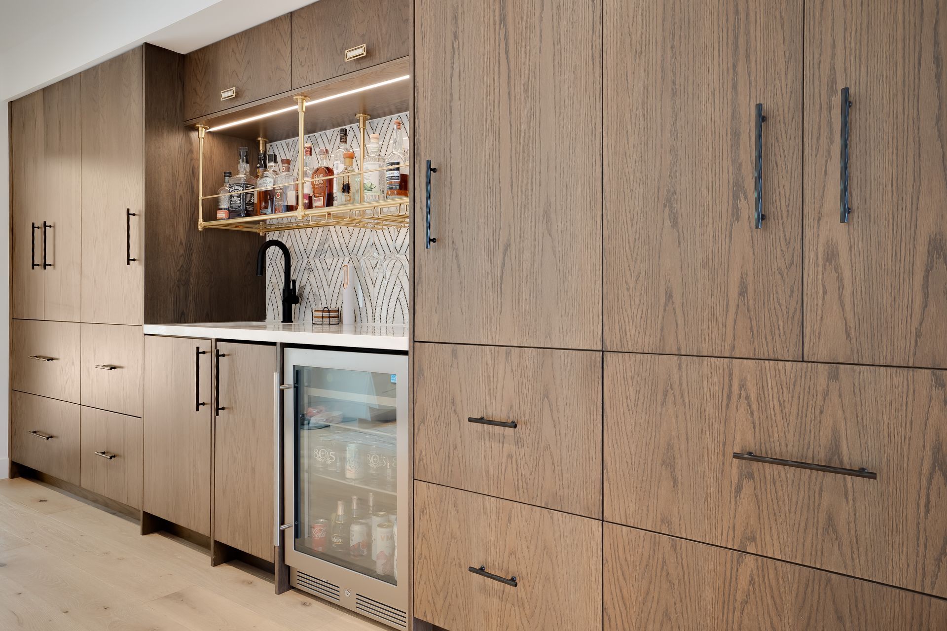 Built-in wooden bar with liquor bottles, mini-fridge, and a backsplash, with dark hardware and lighting.