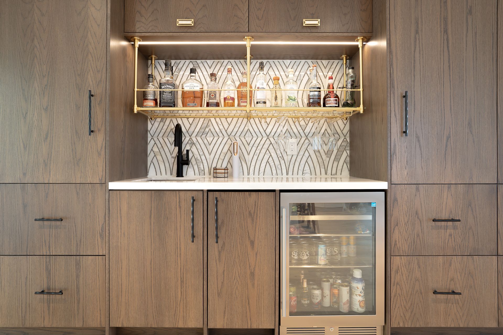 Built-in home bar with wooden cabinets, gold shelving, and a mini-fridge, with liquor bottles.