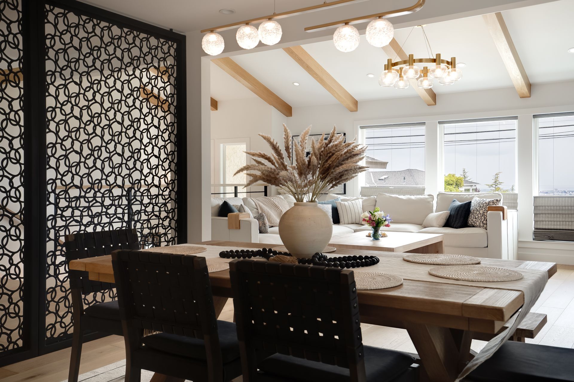 Dining area with wooden table, chairs, and decorative screen in front of a living room with a white sofa and large windows.
