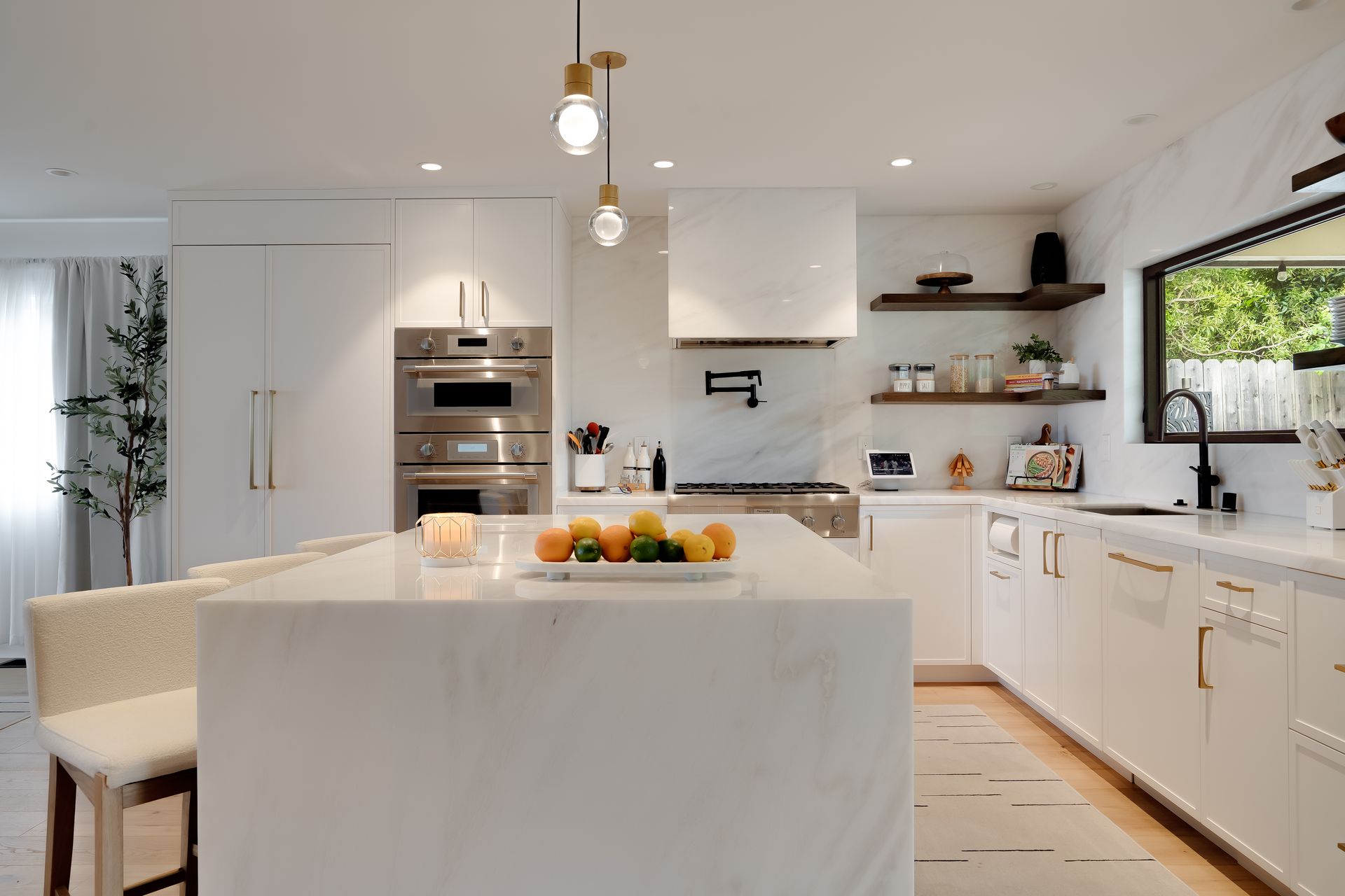 Modern white kitchen with island, oven, and wooden shelves; fruit on island.