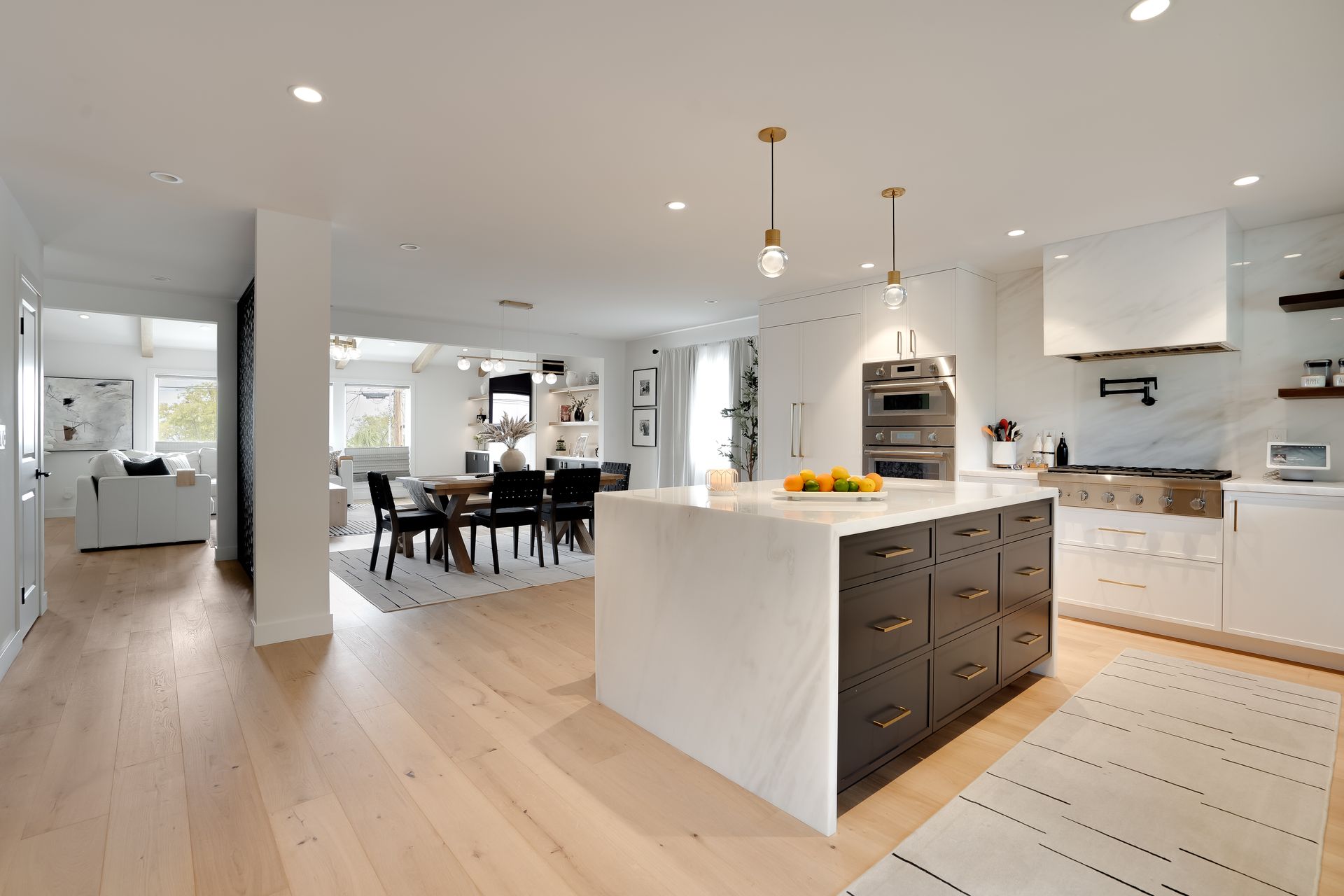Modern kitchen with white cabinets, marble island, and light wood floors. Dining area and living room in background.