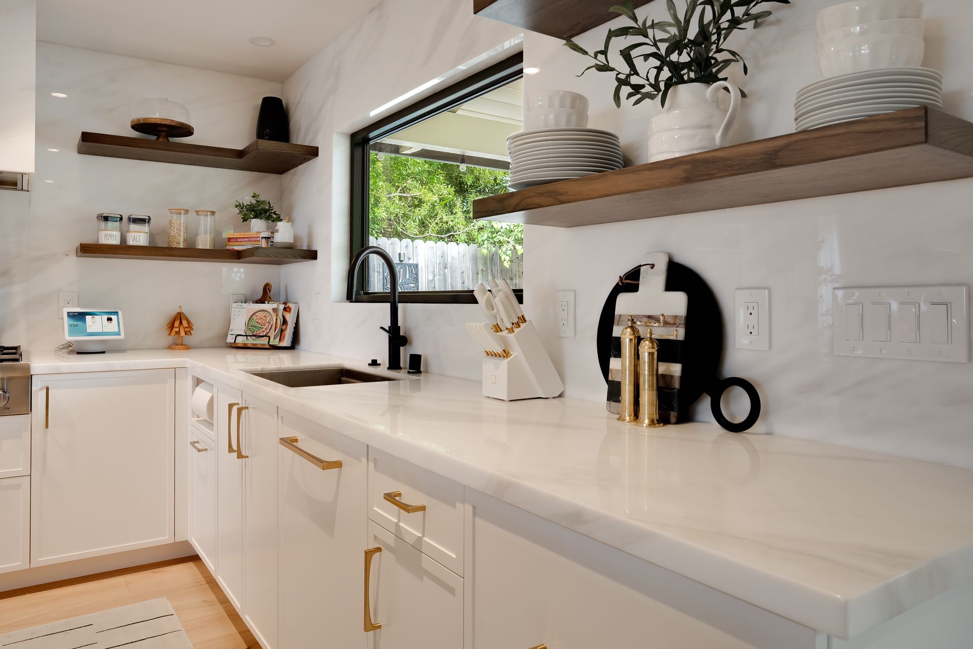 White kitchen with marble countertops, open shelves, and a window overlooking greenery.