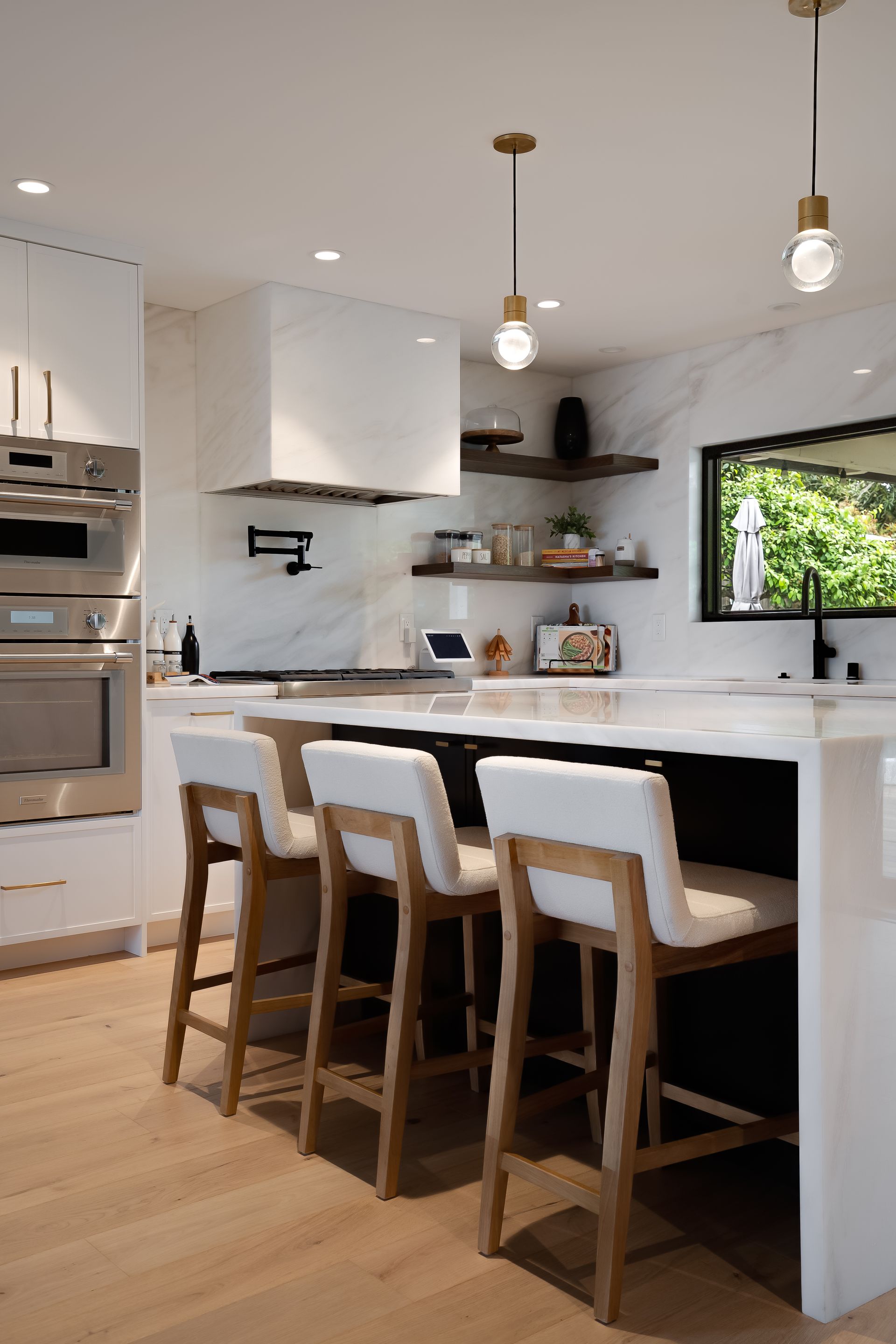 Modern kitchen with white island, stools, and range hood. Light wood floors, stainless steel appliances.