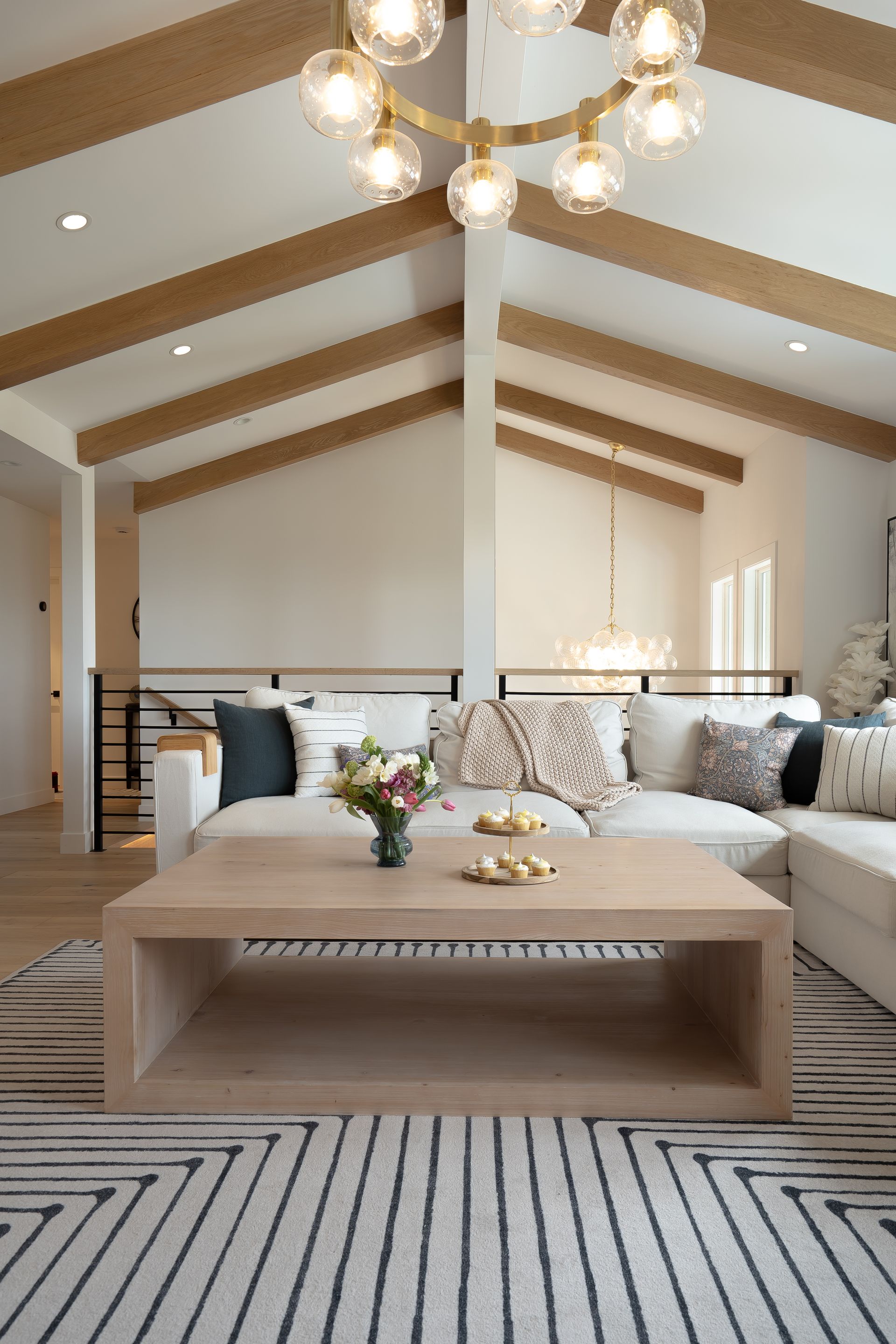 Living room with light wood beams, white walls, sectional sofa, patterned rug, and a modern chandelier.