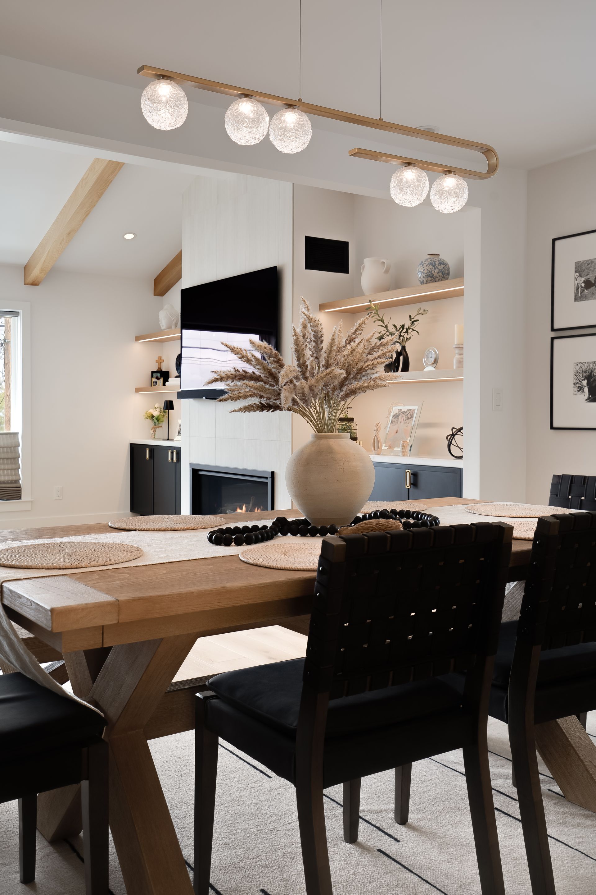 Dining room with wooden table, black chairs, and a light fixture. Fireplace and shelves in the background.