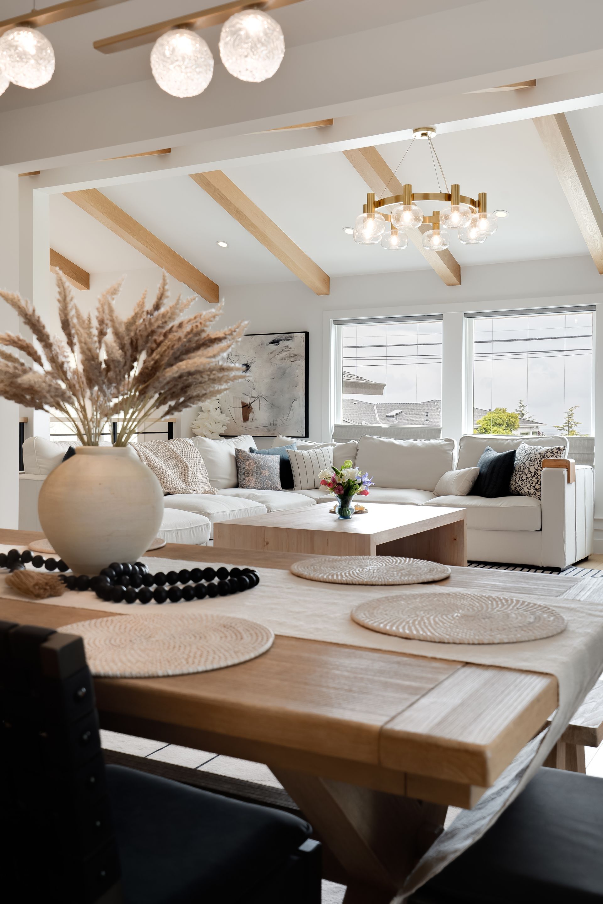 Dining table set for a meal, leading into a bright living room with white couches and a chandelier.
