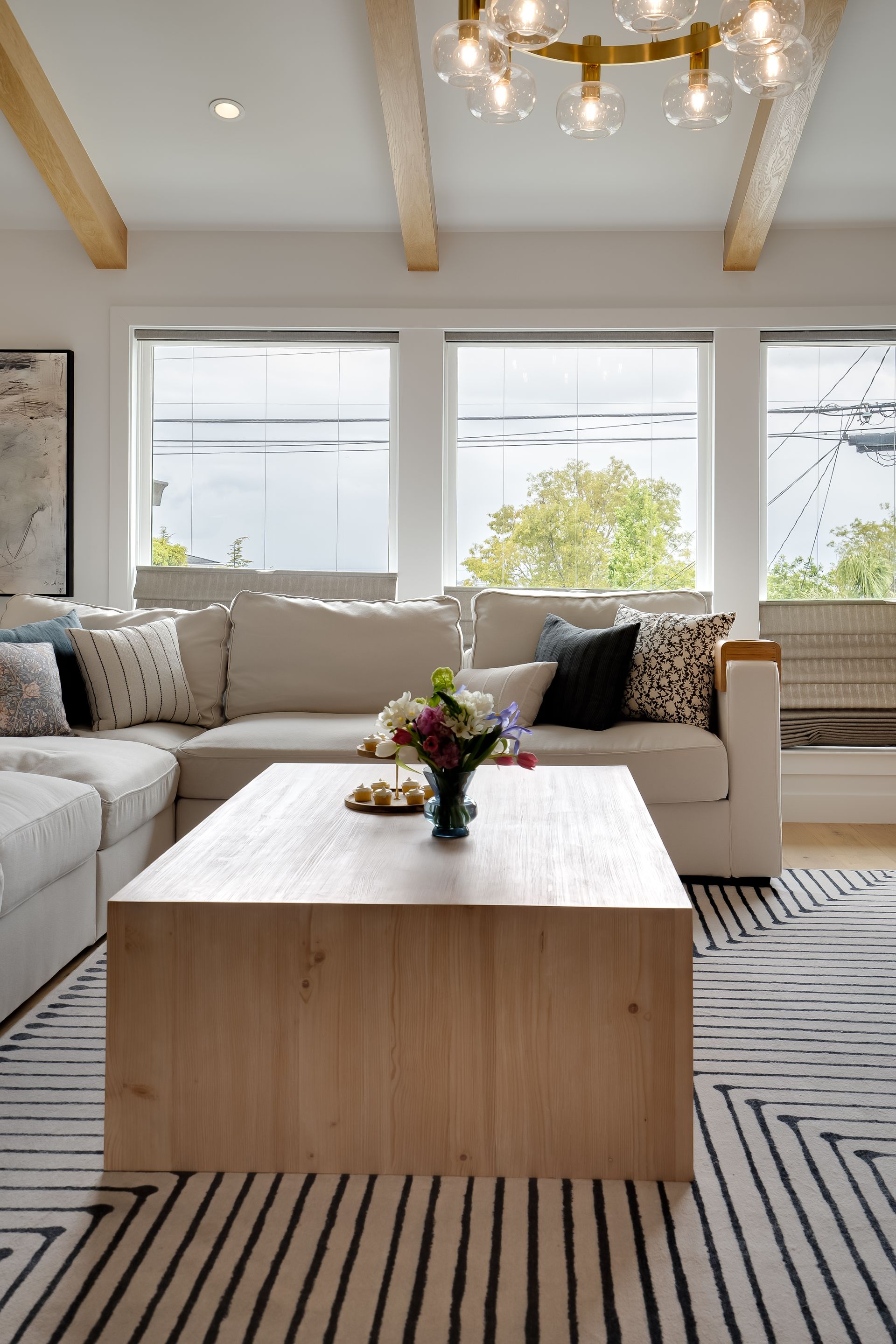 Living room with neutral sectional, wooden coffee table, and patterned rug. Large windows and a decorative chandelier.