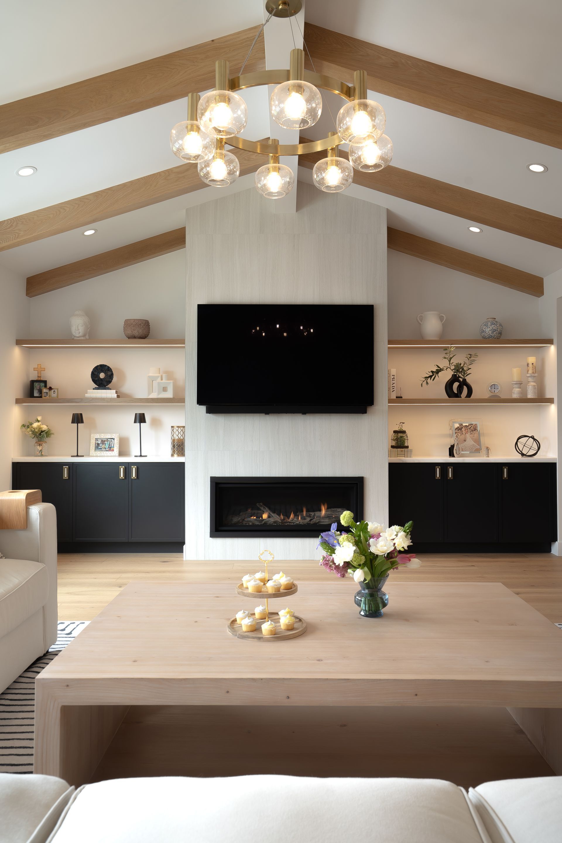 Living room with fireplace, TV, built-in shelves, light wood beams, and gold chandelier.