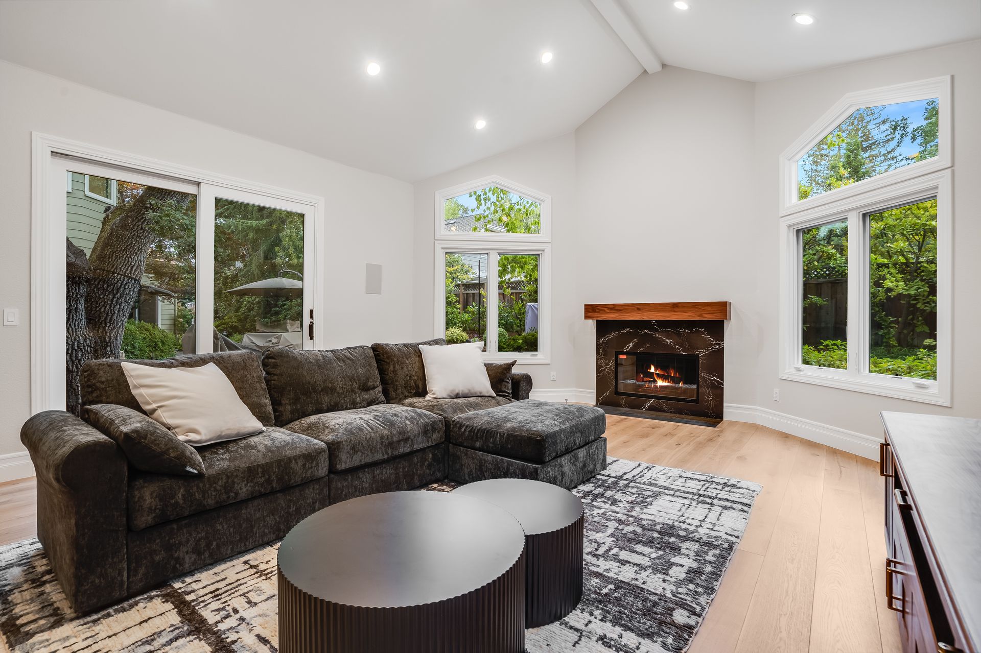Spacious living room with dark sectional, fireplace, and large windows overlooking greenery.
