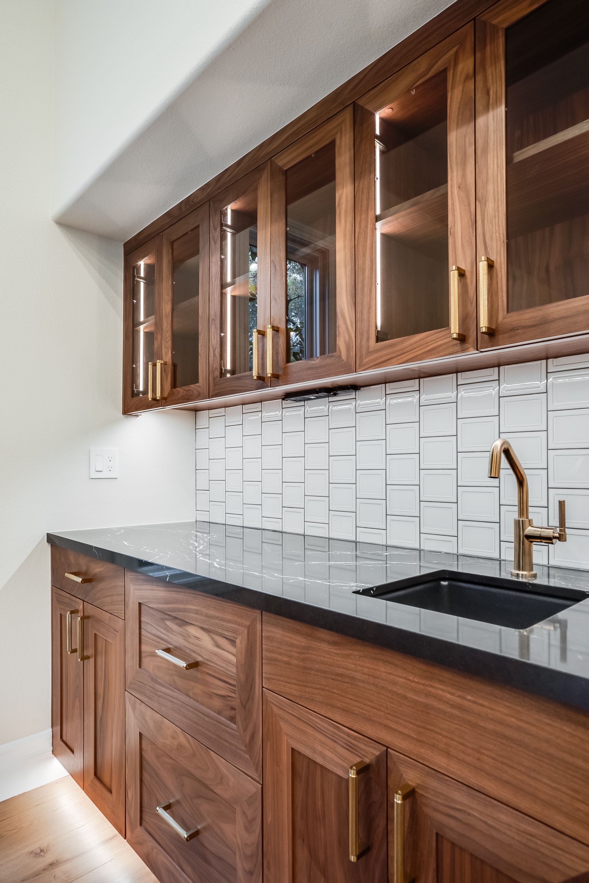 Built-in wet bar with wood cabinets, glass-front uppers, black countertop, gold faucet, and white tile backsplash.