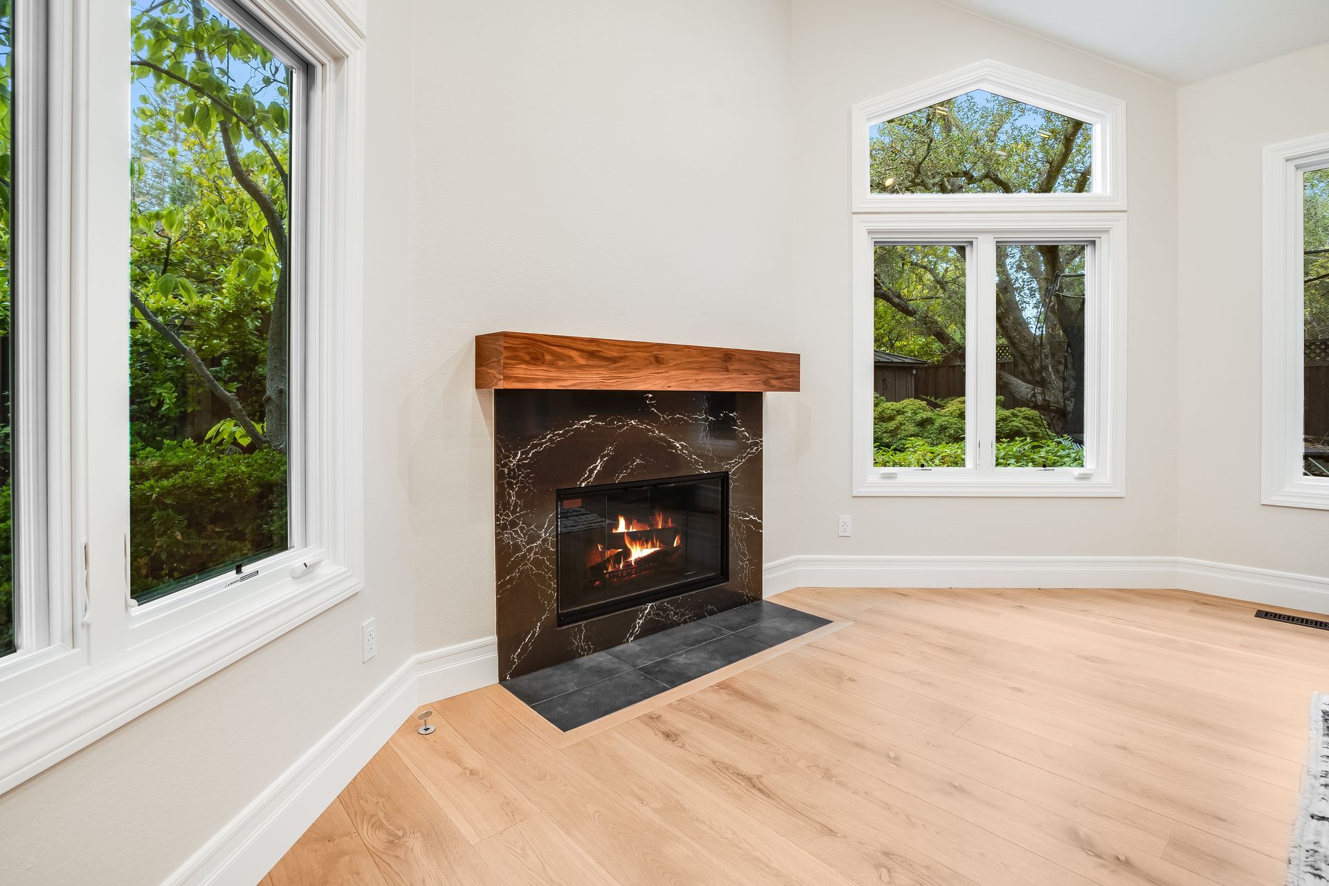Fireplace with a black marble surround and wooden mantel, flanked by windows overlooking greenery.