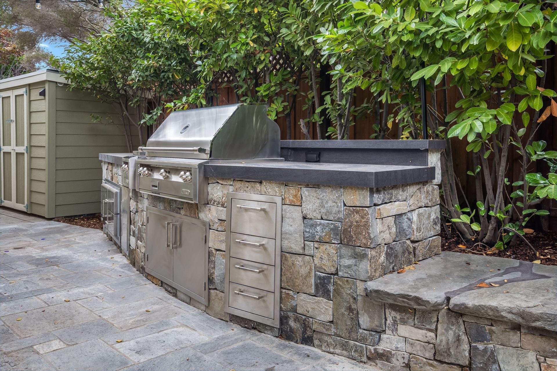 Outdoor kitchen with stainless steel grill and stone counter, next to a shed and greenery.