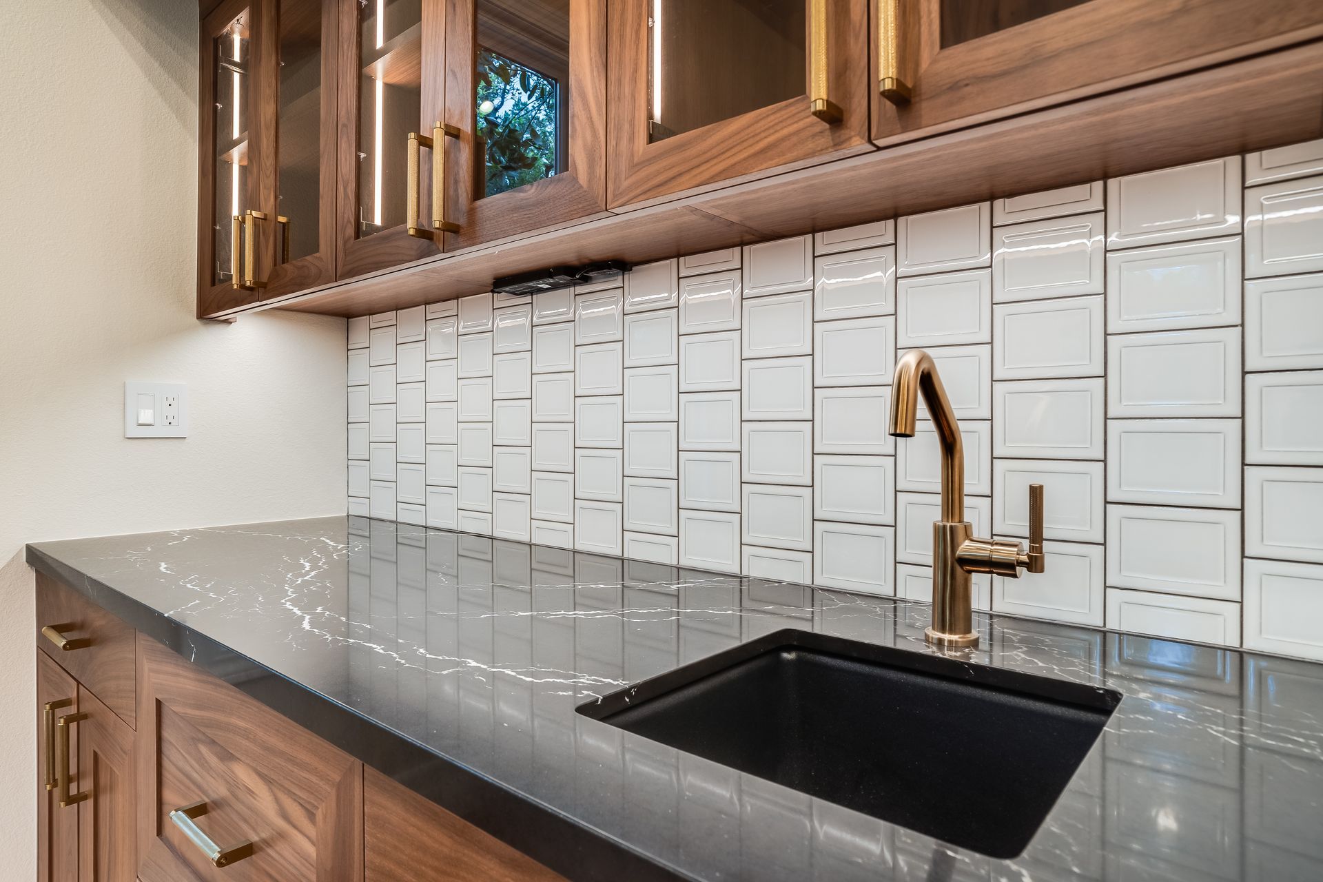Dark cabinetry with gold hardware, white tiled backsplash, black sink, and gold faucet.