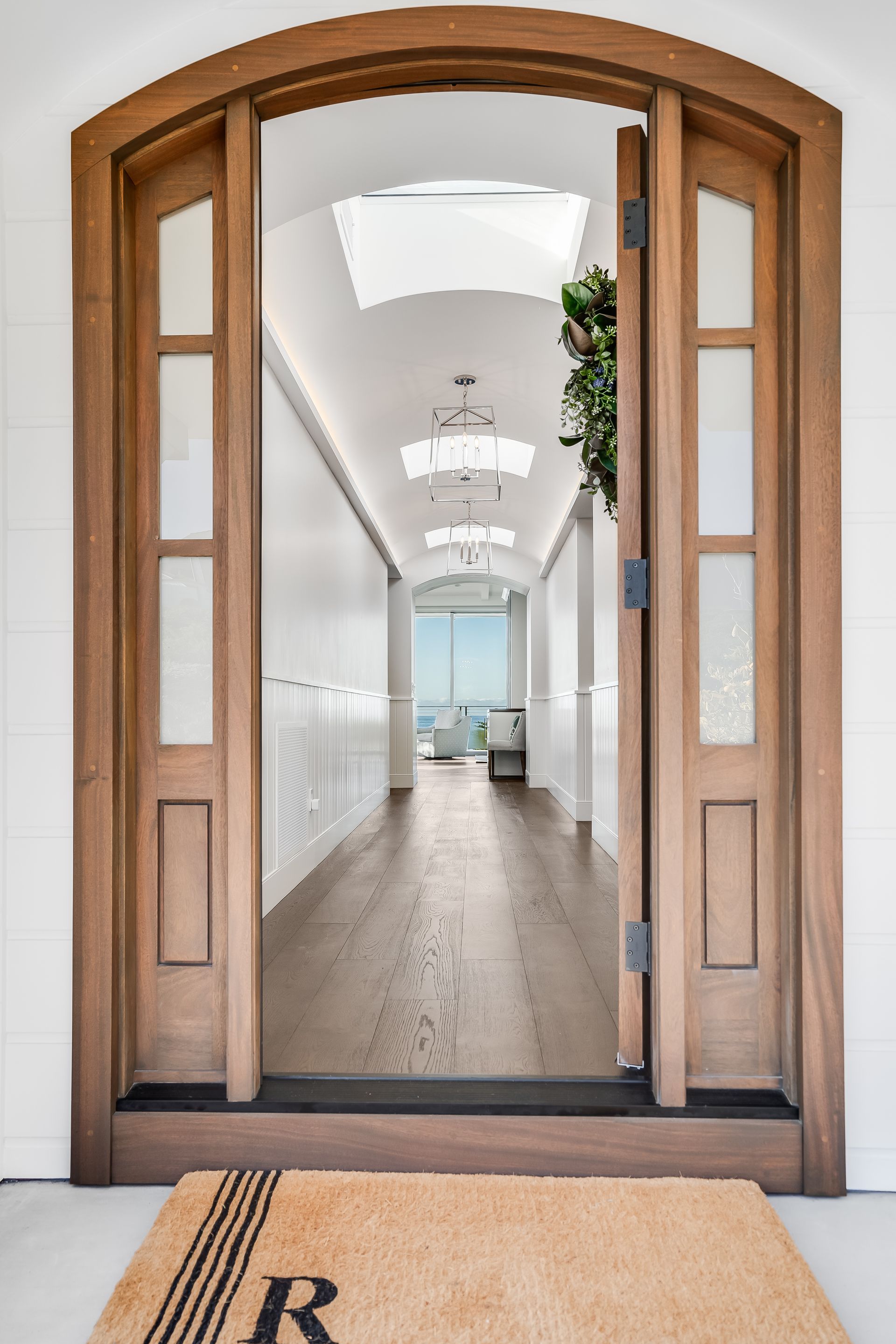Wooden doorway open to a hallway with white walls, wood floor, and ocean view; doormat reads