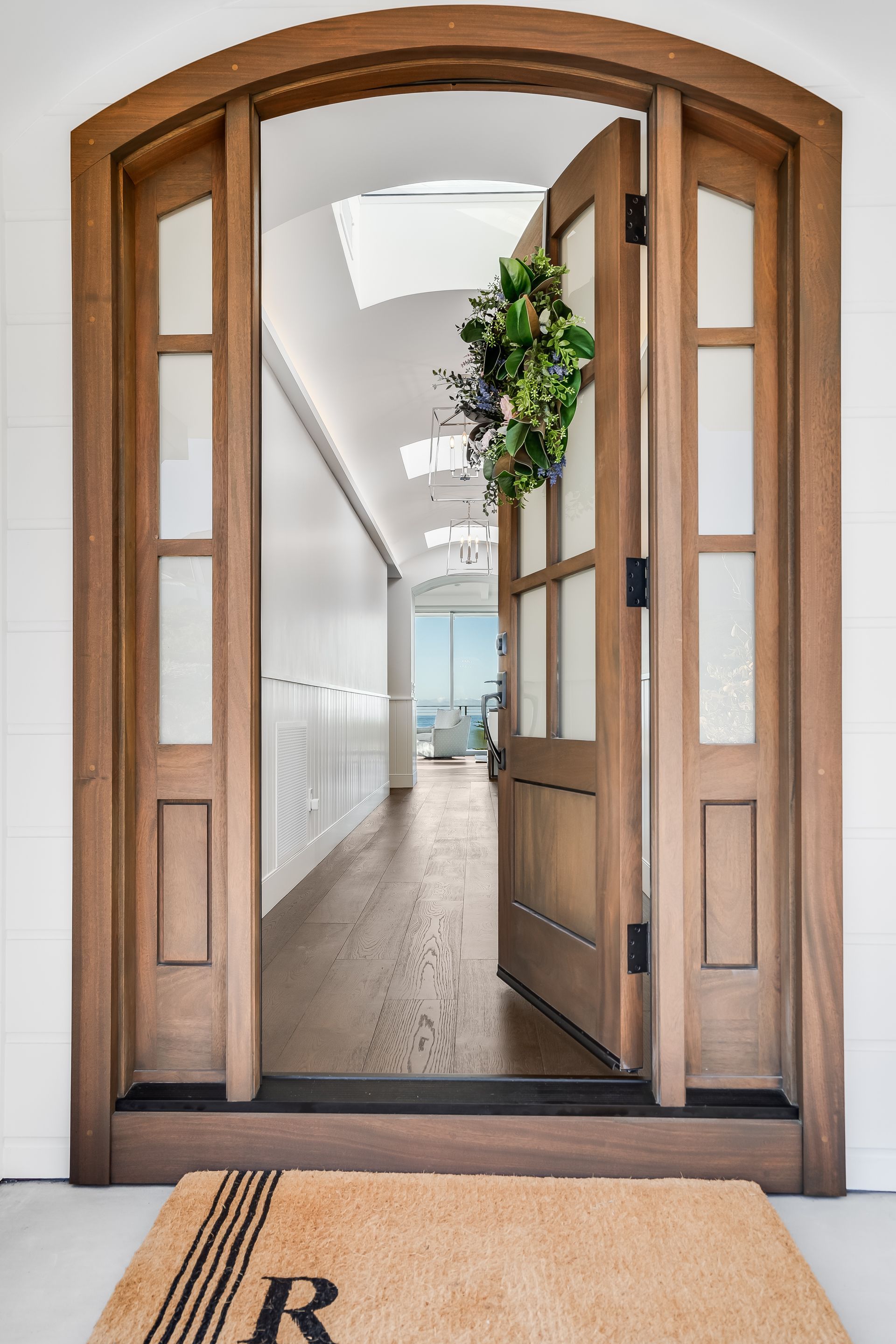 Open wooden doorway with wreath, leading to hallway. Brown door with glass panes, tan doormat, white walls.