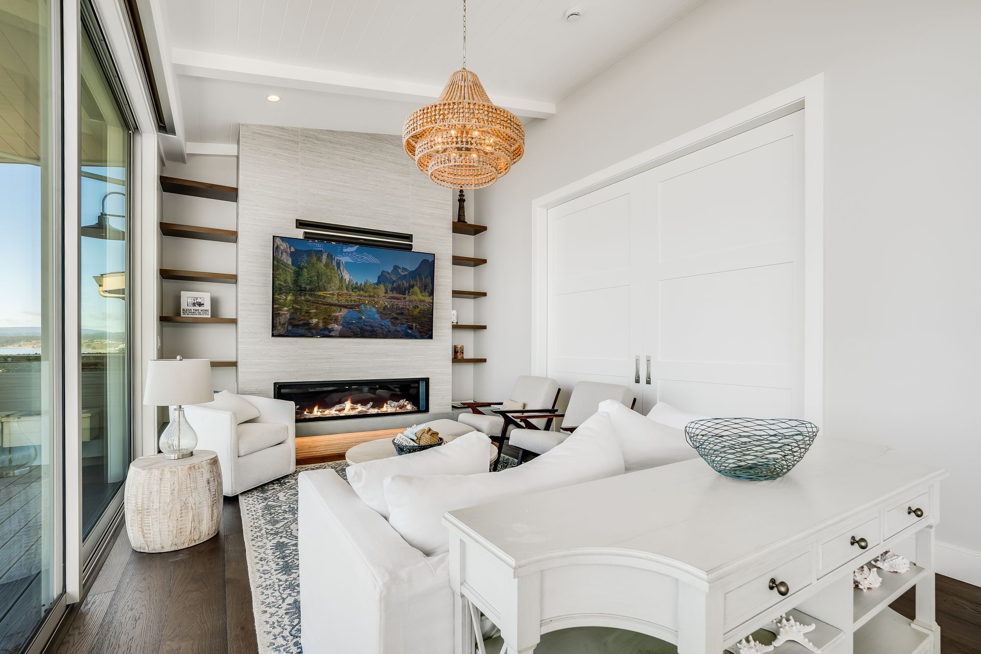 Bright living room with white furniture, fireplace, shelves, and large windows.