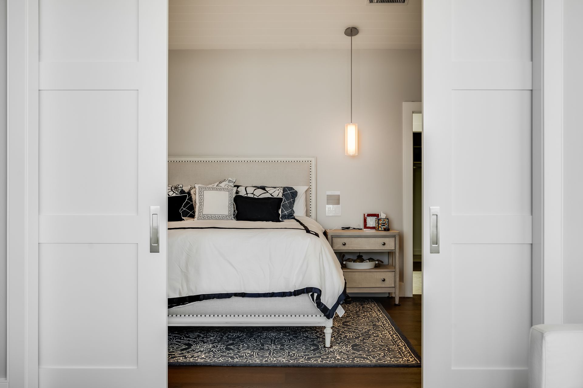 White bedroom interior visible through open sliding doors. Bed with white comforter, nightstand, and area rug.