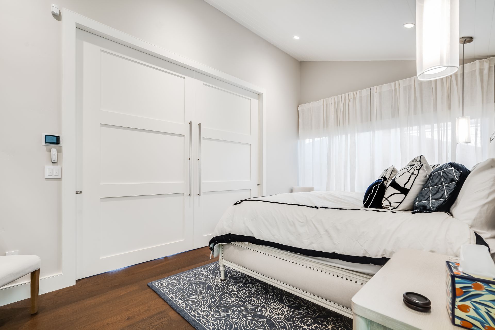 White bedroom with closed double doors, bed, rug, and sheer curtains.