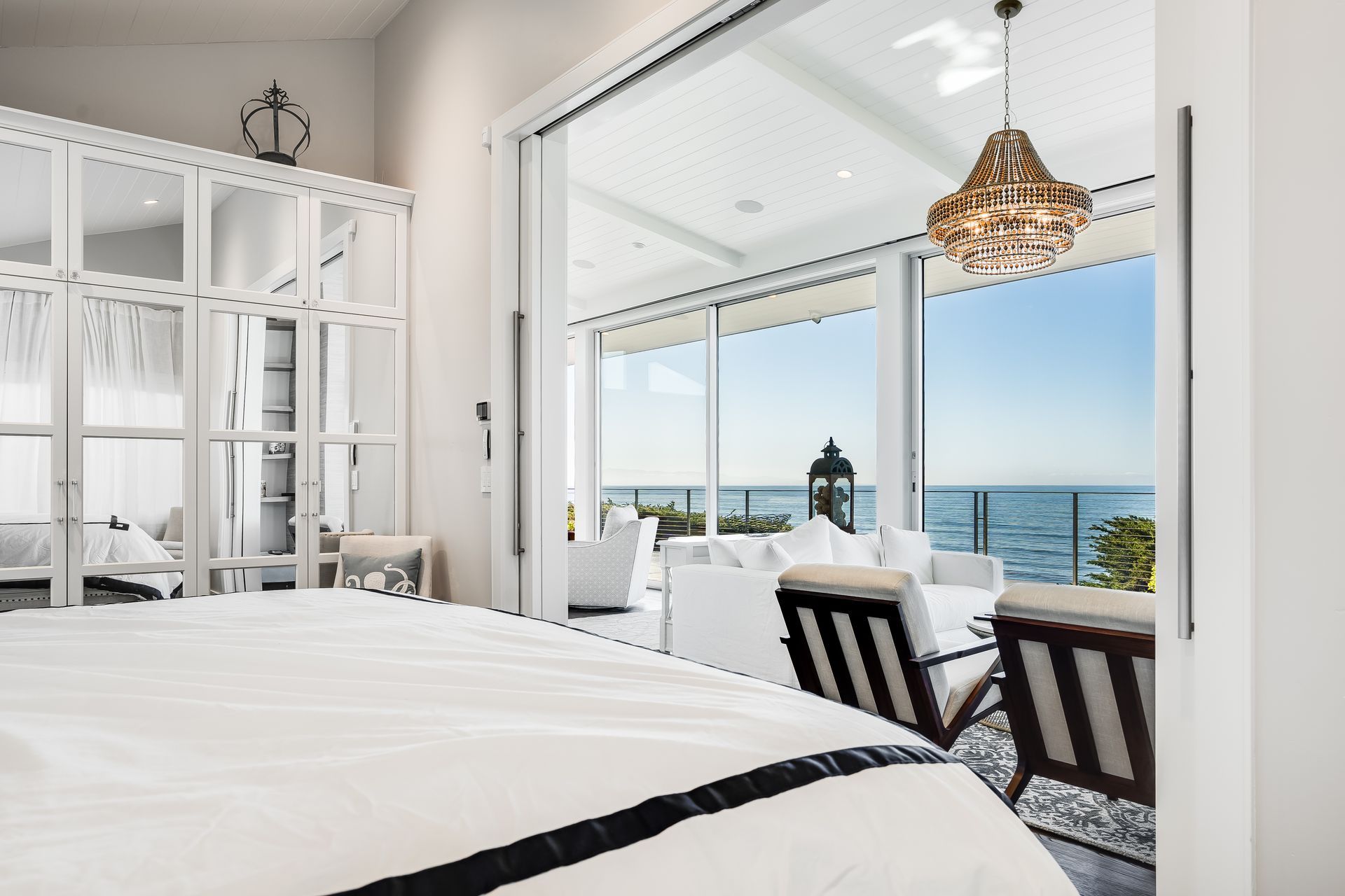 Bedroom with a white bed, large windows opening to a patio with ocean view and chairs.