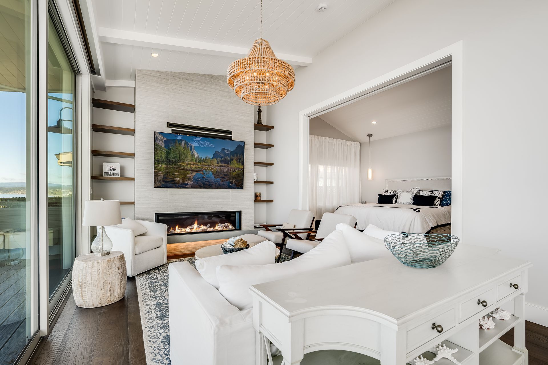 Cozy white living room with fireplace, TV, chandelier, and a view into a bedroom.