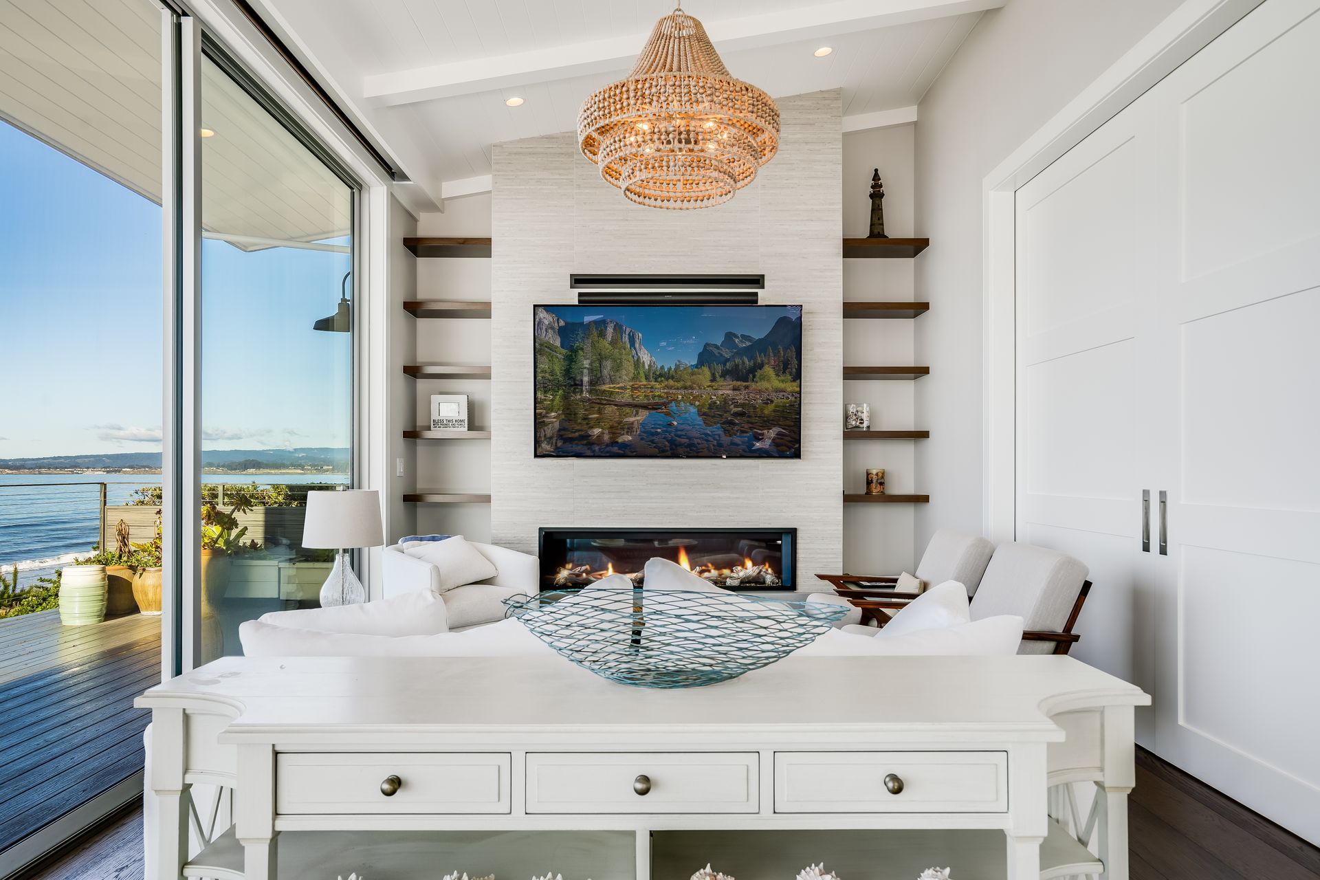 Coastal living room with large windows, fireplace, built-in shelves, and white furniture.