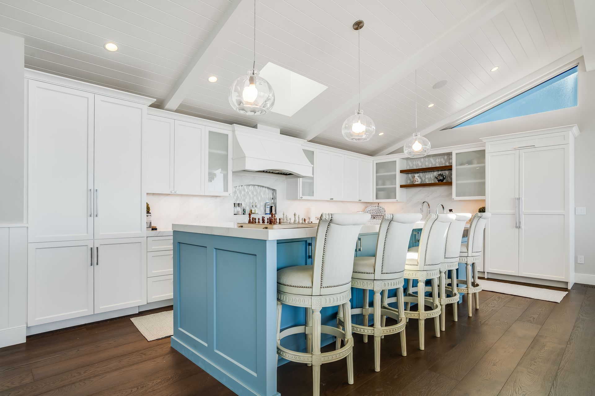 Bright kitchen with a blue island, white cabinets, and wooden floor. Chairs at island, pendant lights.