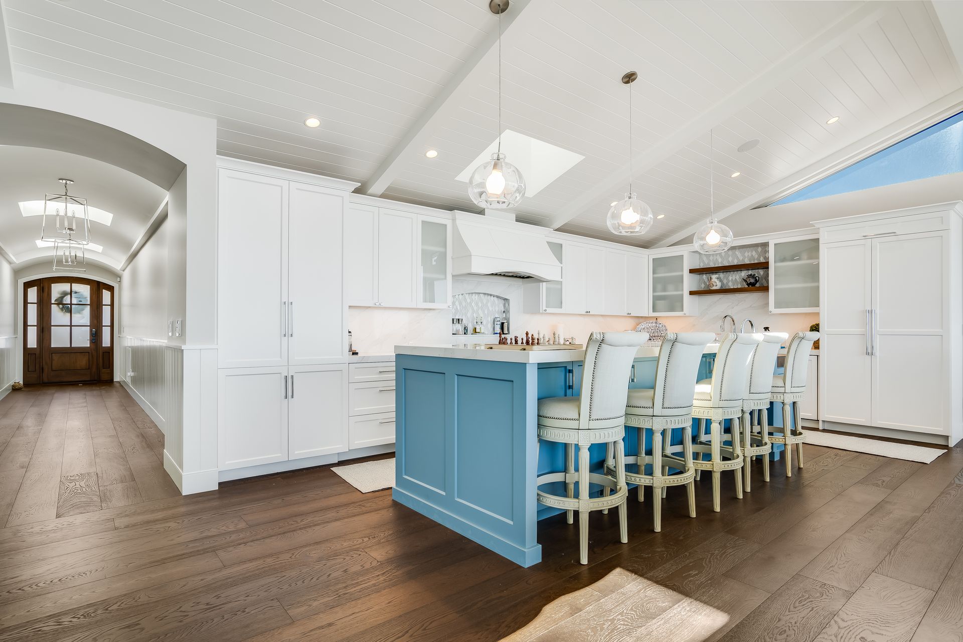 Spacious white kitchen with blue island, wooden floor, and seating. Hallway with a wooden door.
