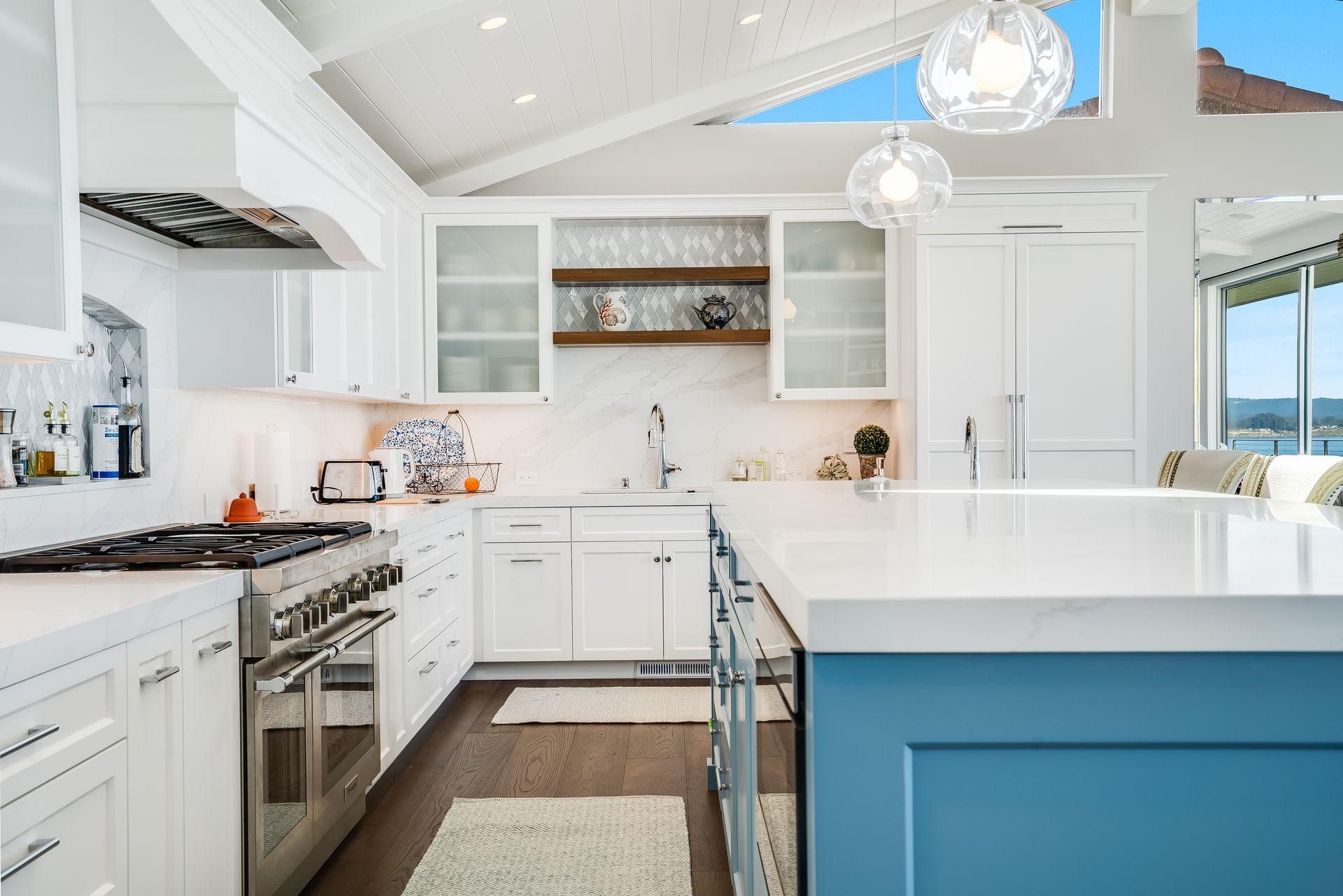 Bright white kitchen with a blue island, stainless steel appliances, and a view.