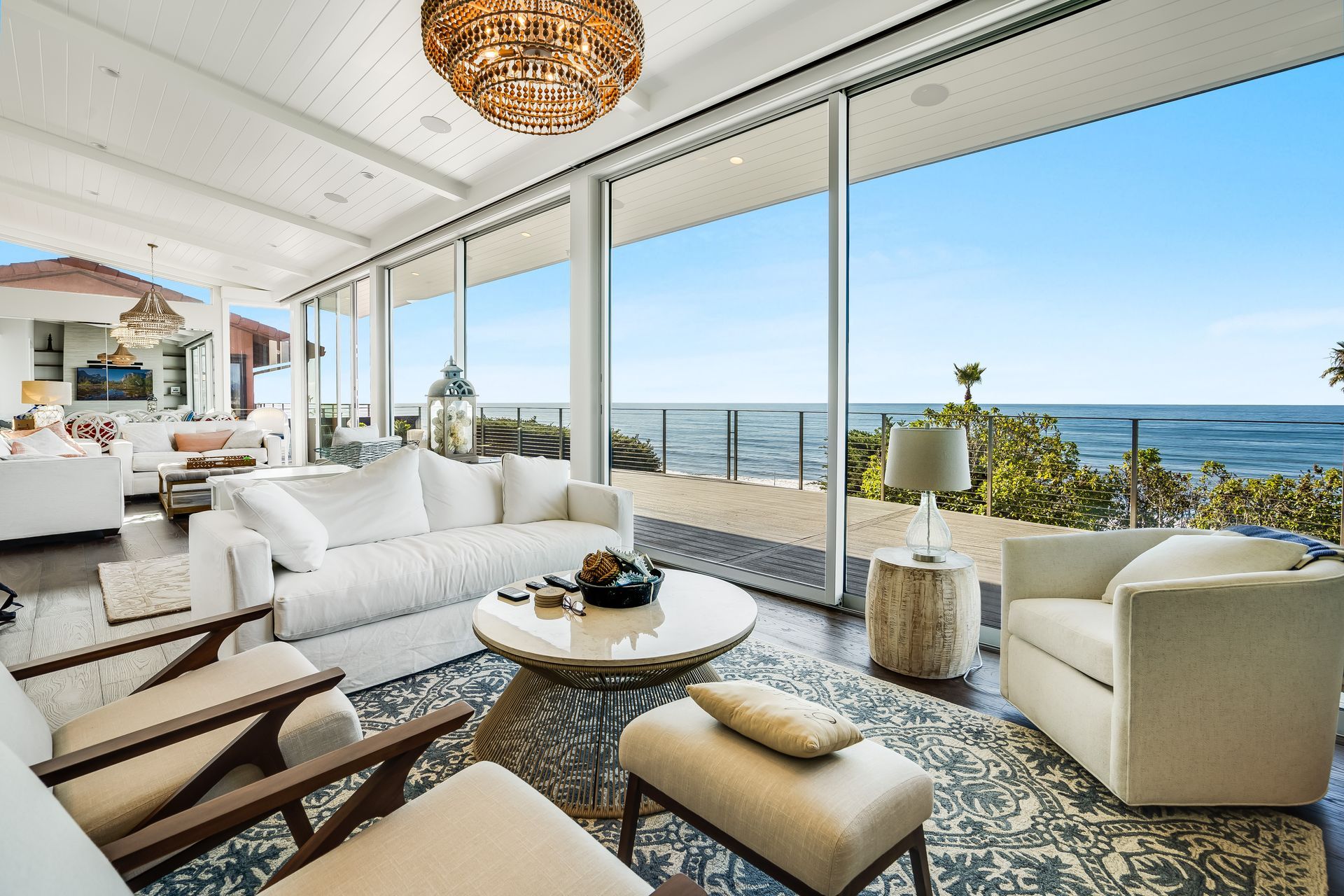 Living room with ocean view: white furniture, dark wood floors, large windows, blue sky, and a chandelier.