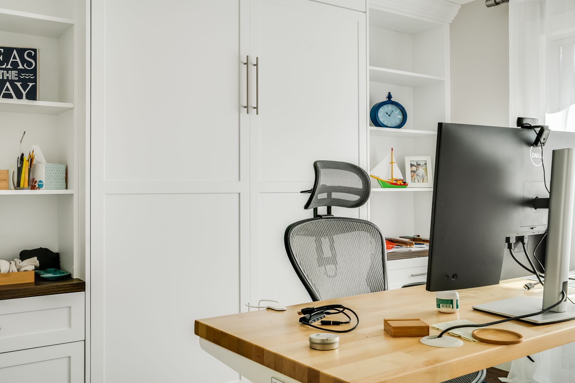 Home office with white cabinets, desk, and ergonomic chair. A monitor and desk accessories are on the wooden desk.