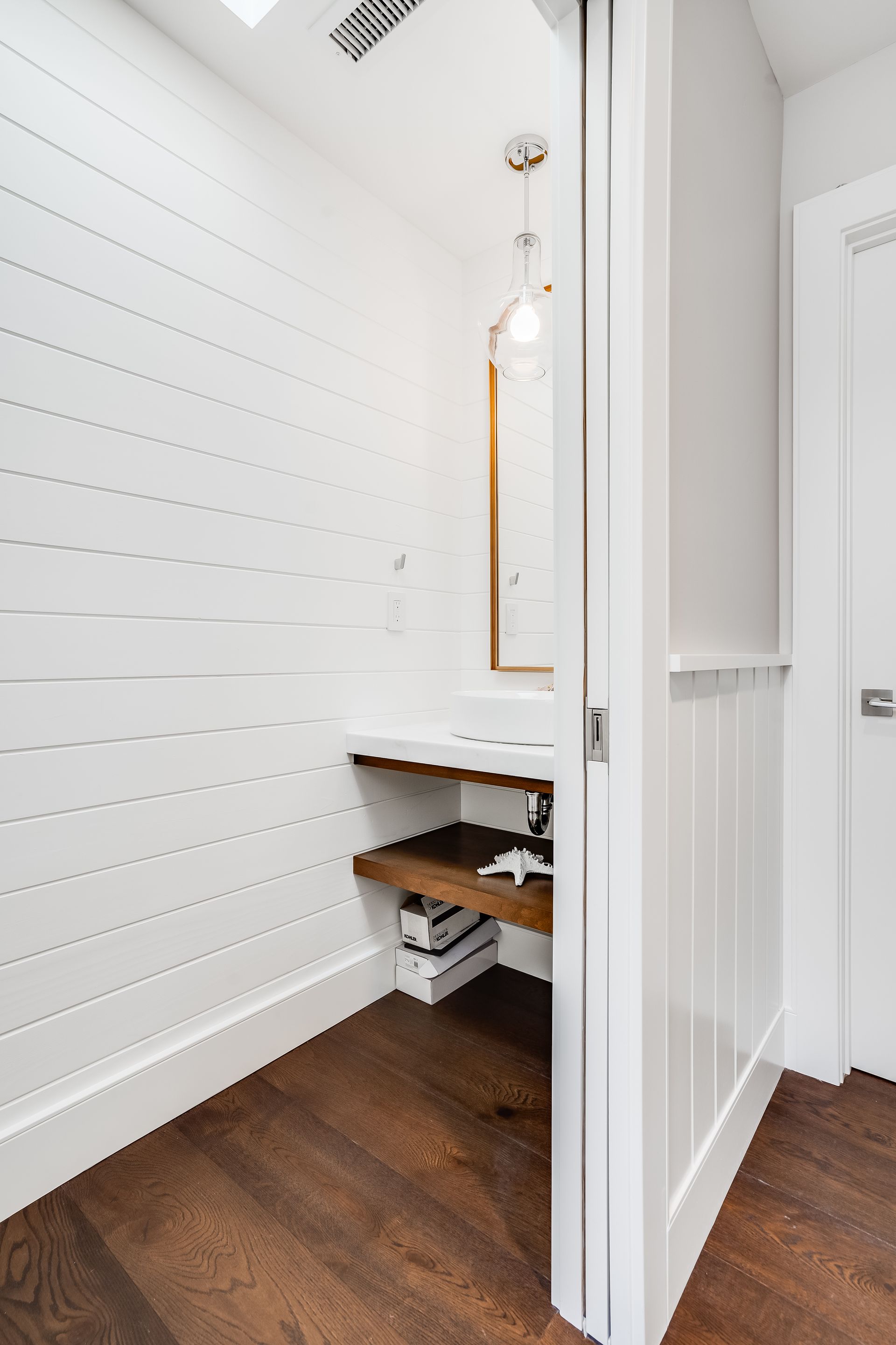 Small, white-walled bathroom nook with wooden shelves, dark wood floor, and a hanging light fixture.