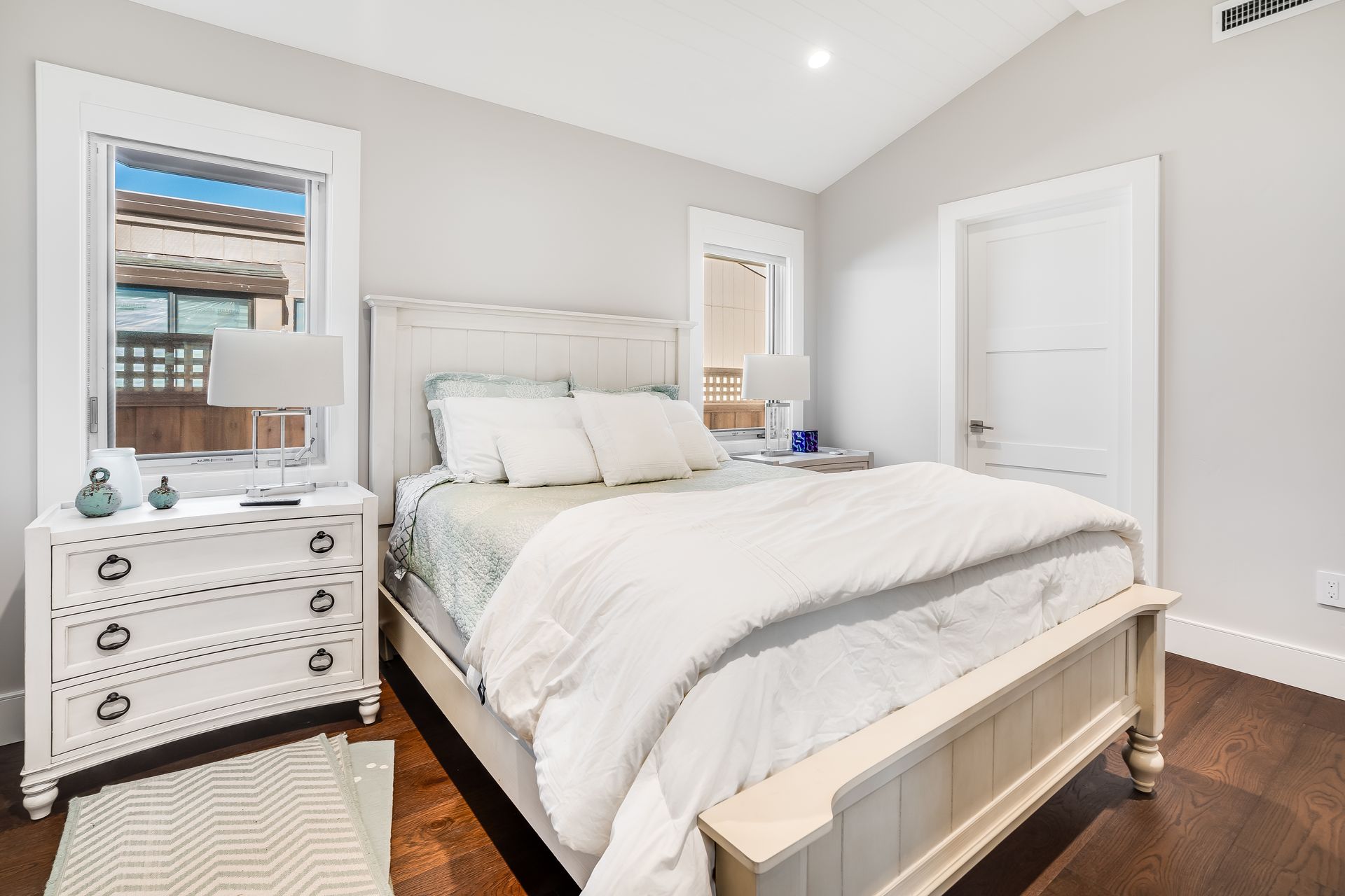 White bedroom with bed, nightstand, window, and door. Dark wood floor and gray walls.