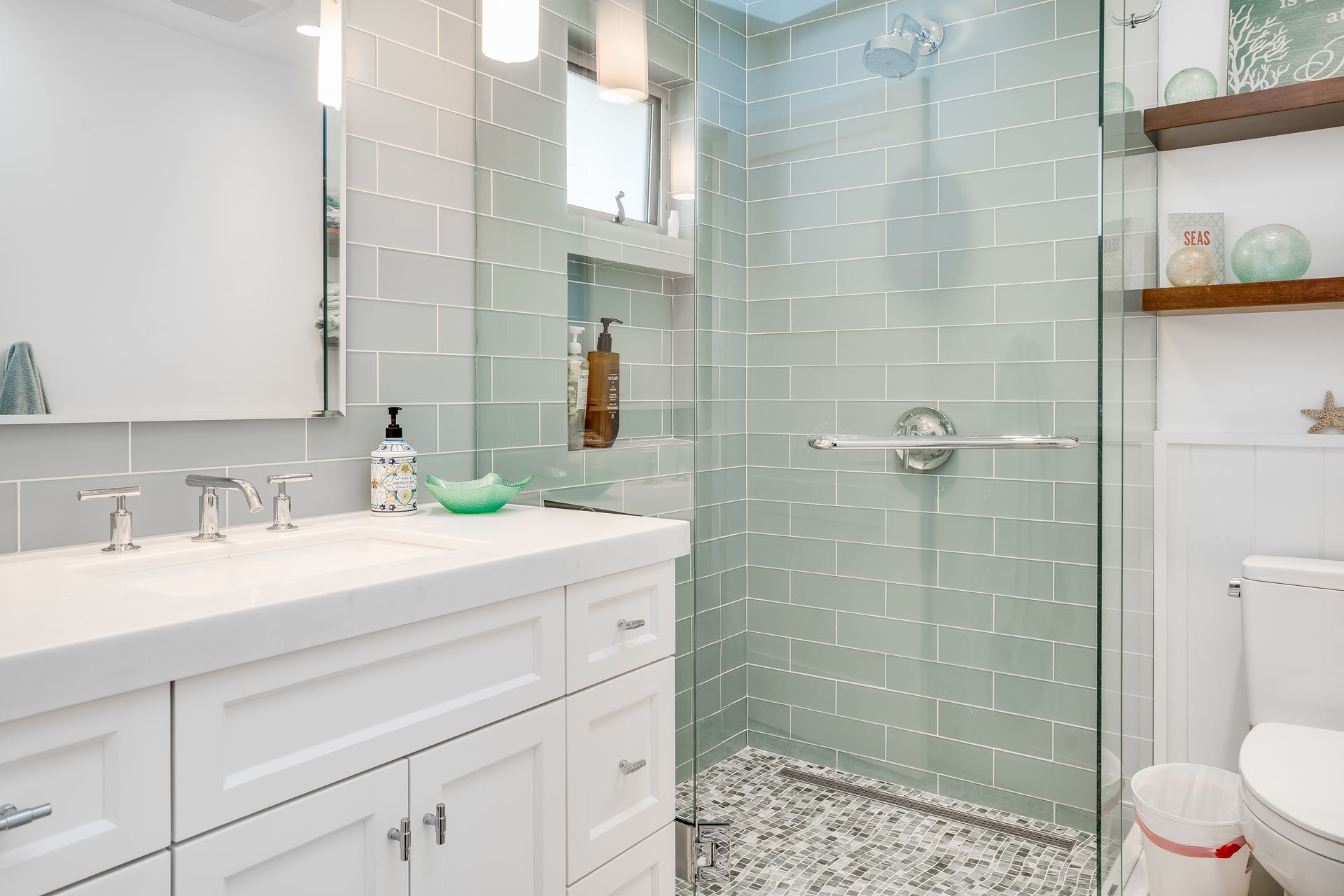 A bright white bathroom with light green tiled shower, white vanity, and a toilet.