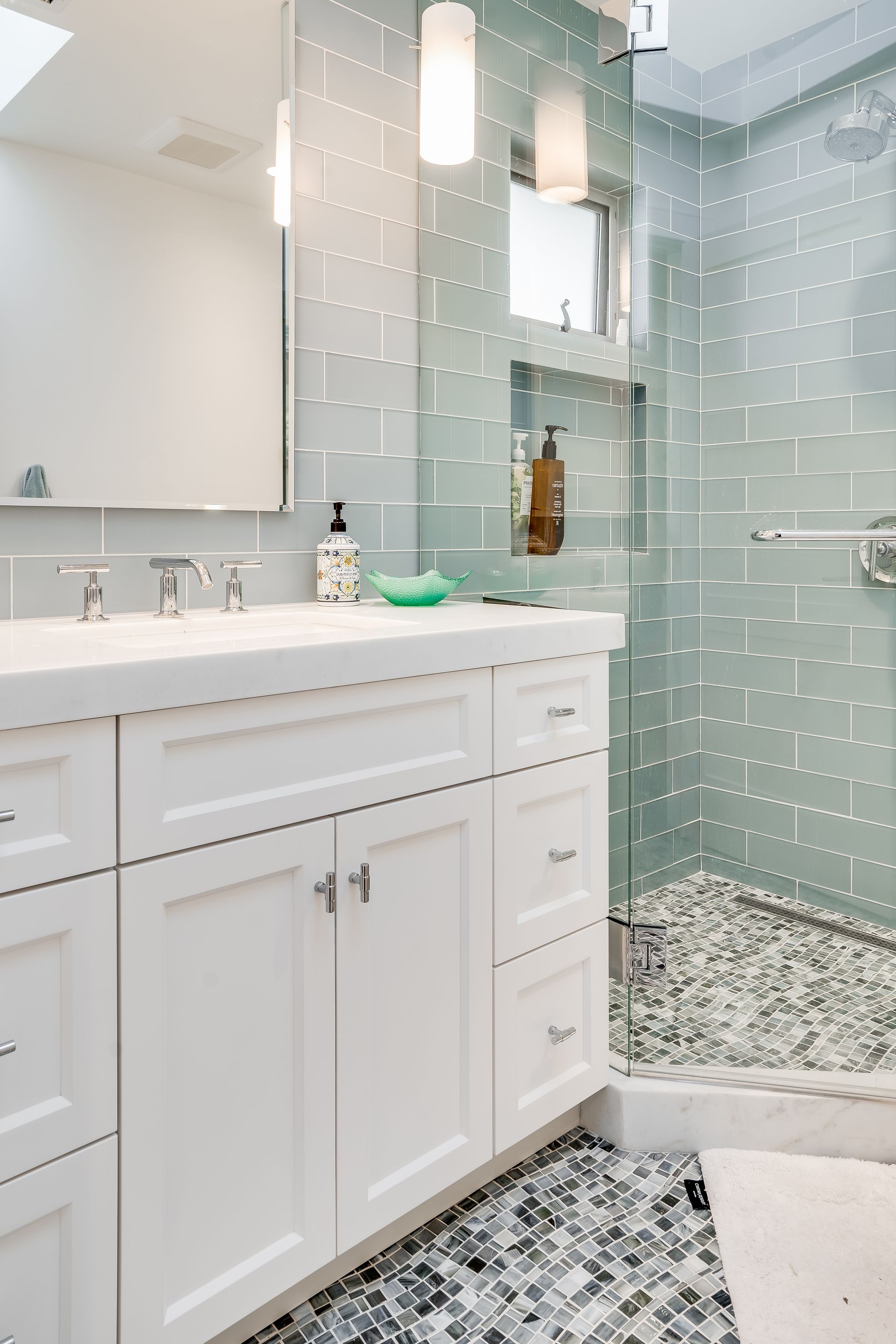 White bathroom with blue-tiled walls, glass shower, pebble floor, white vanity, and bright lighting.