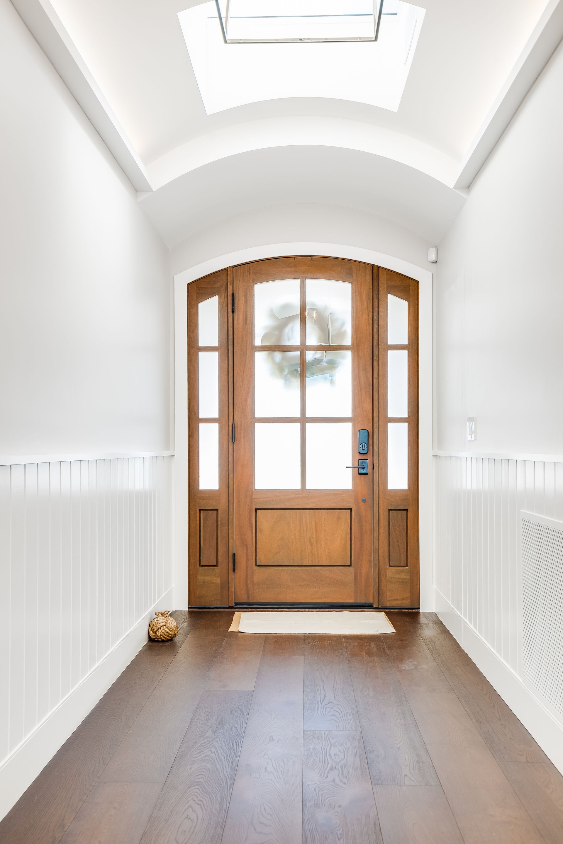 Wooden front door with sidelights and wreath in a white-walled hallway with dark wood floors.
