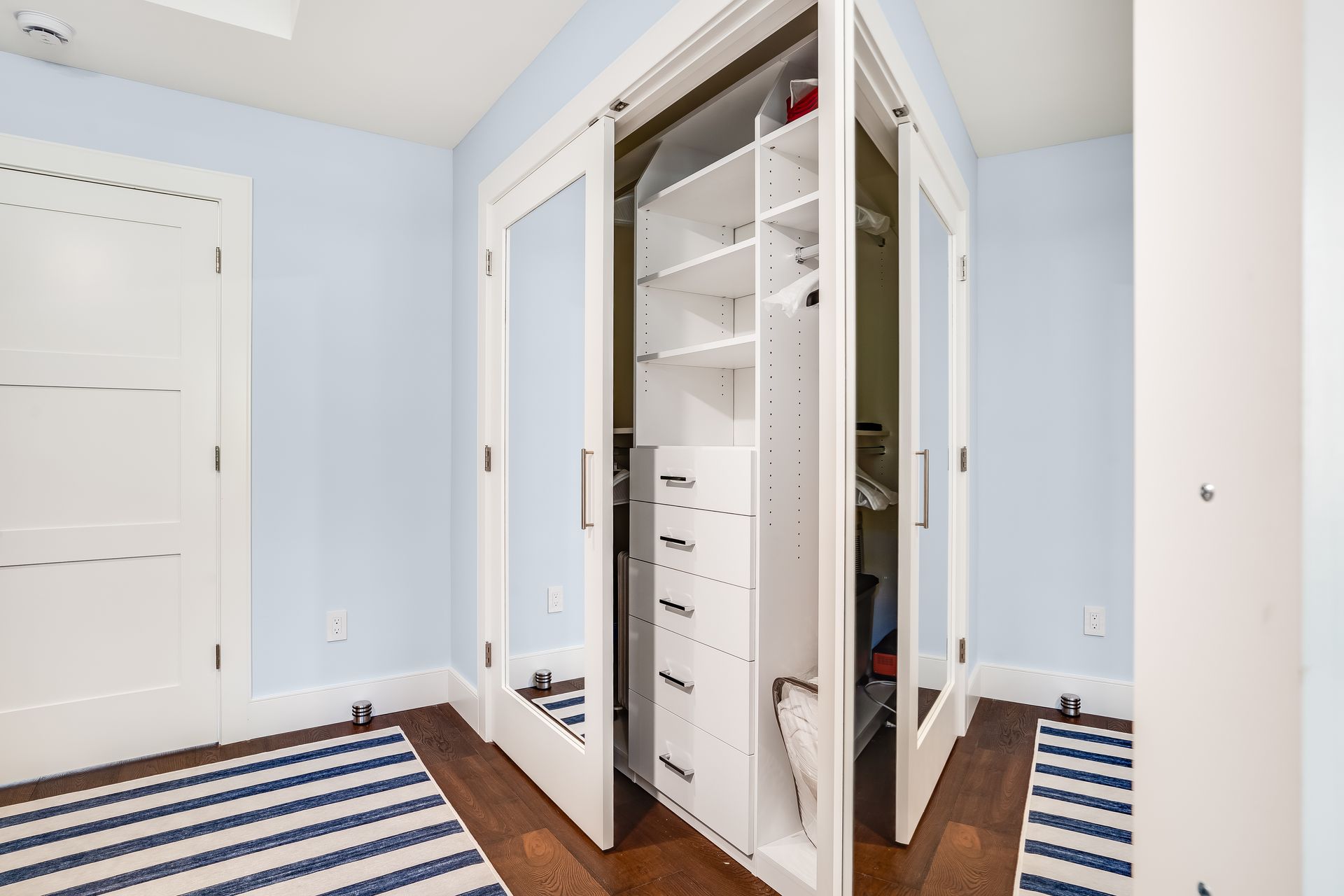 White closet with mirrored doors, corner placement, open to reveal interior shelves and drawers, blue walls, wood floor.