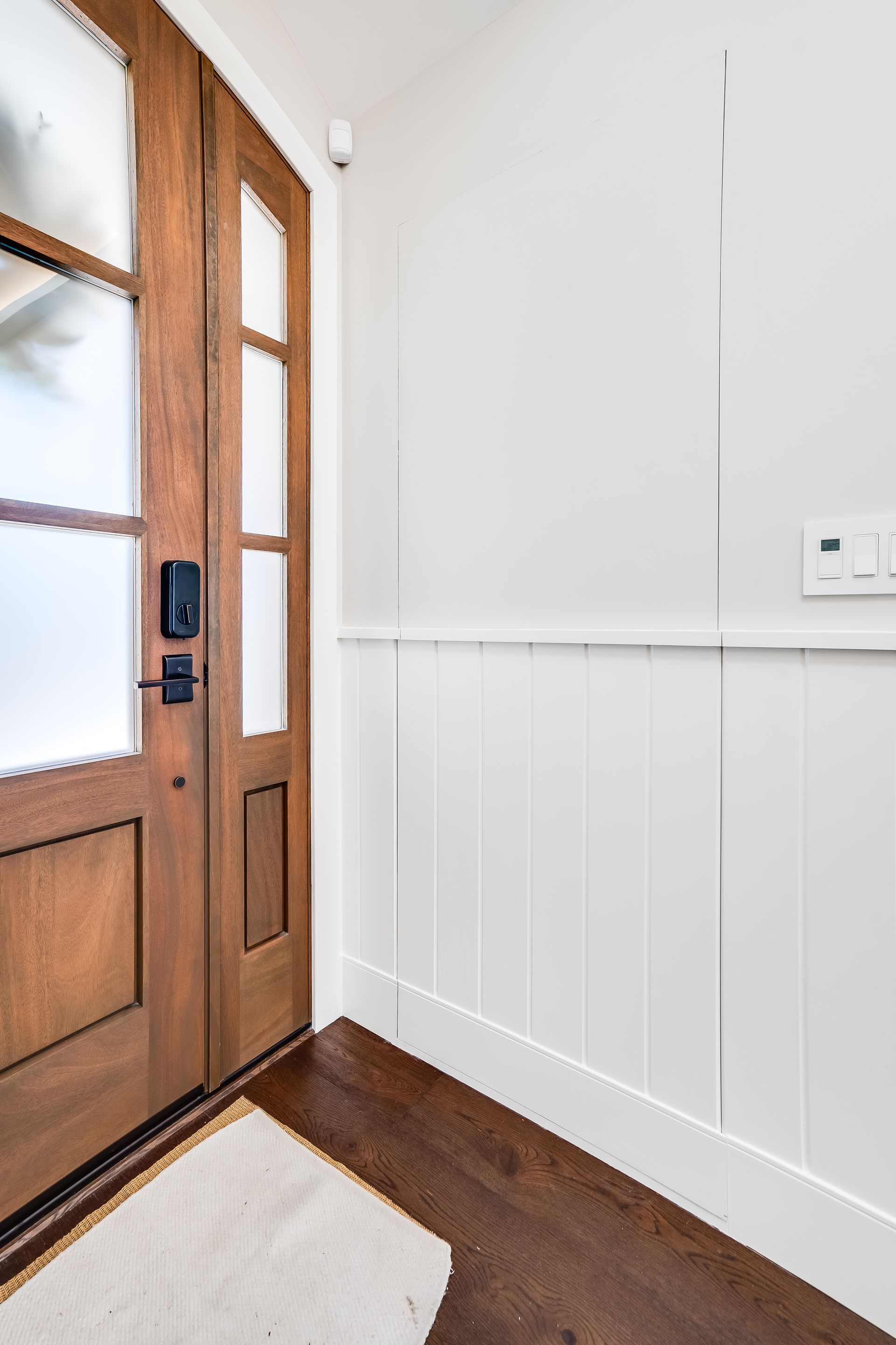 Wooden double front door with frosted glass panels, next to white paneled wall and dark wood floor.