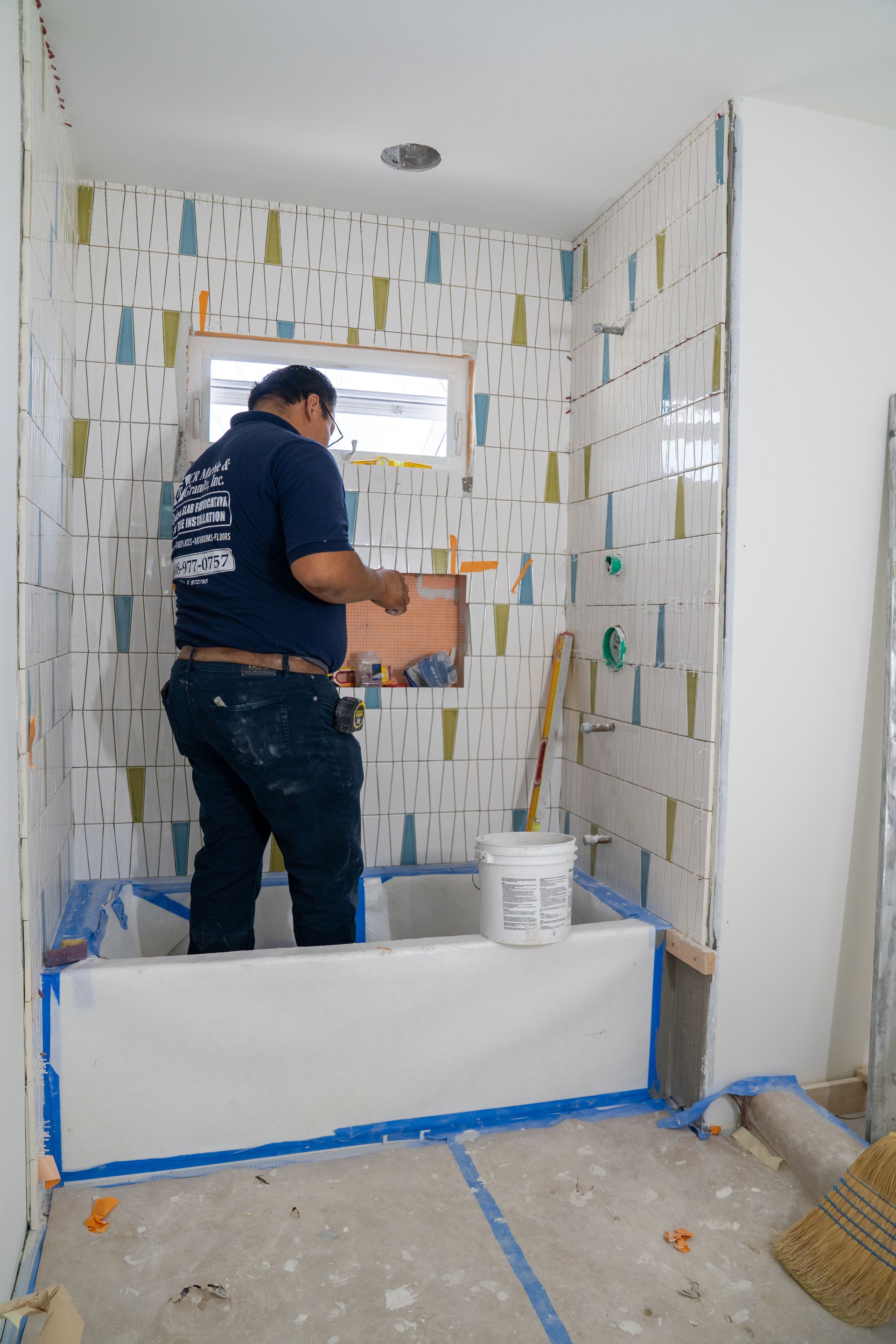 Man tiling a bathroom, standing in tub with colorful accent tile, bucket, window.