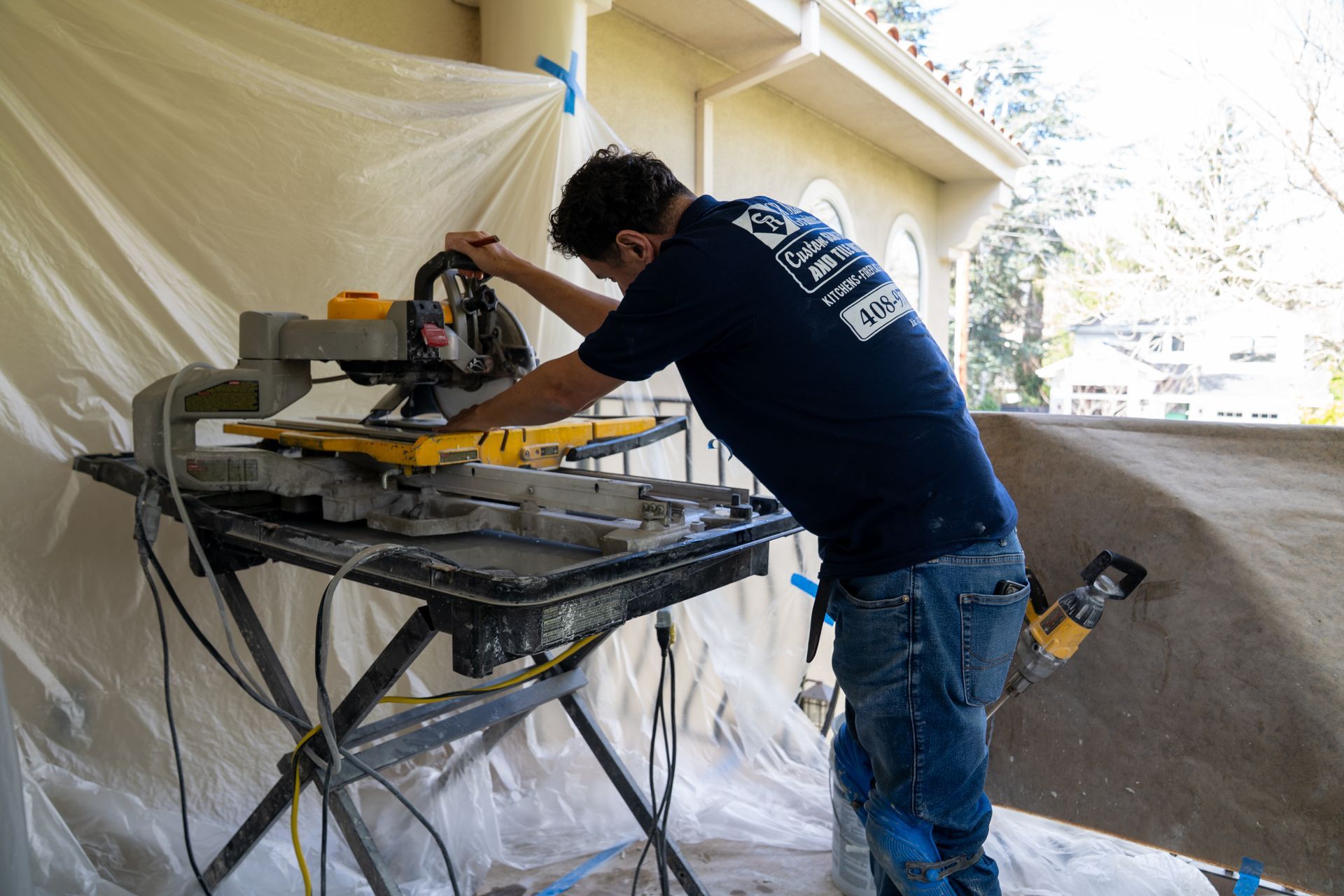 Man in blue shirt using a tile saw outdoors. White protective sheeting covers the area.
