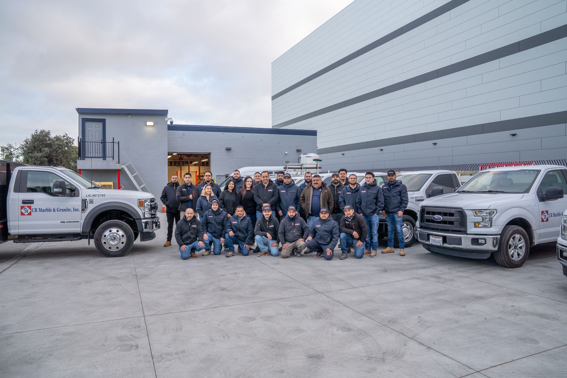 Group of people in front of trucks, building. People are standing, kneeling, and looking at camera.
