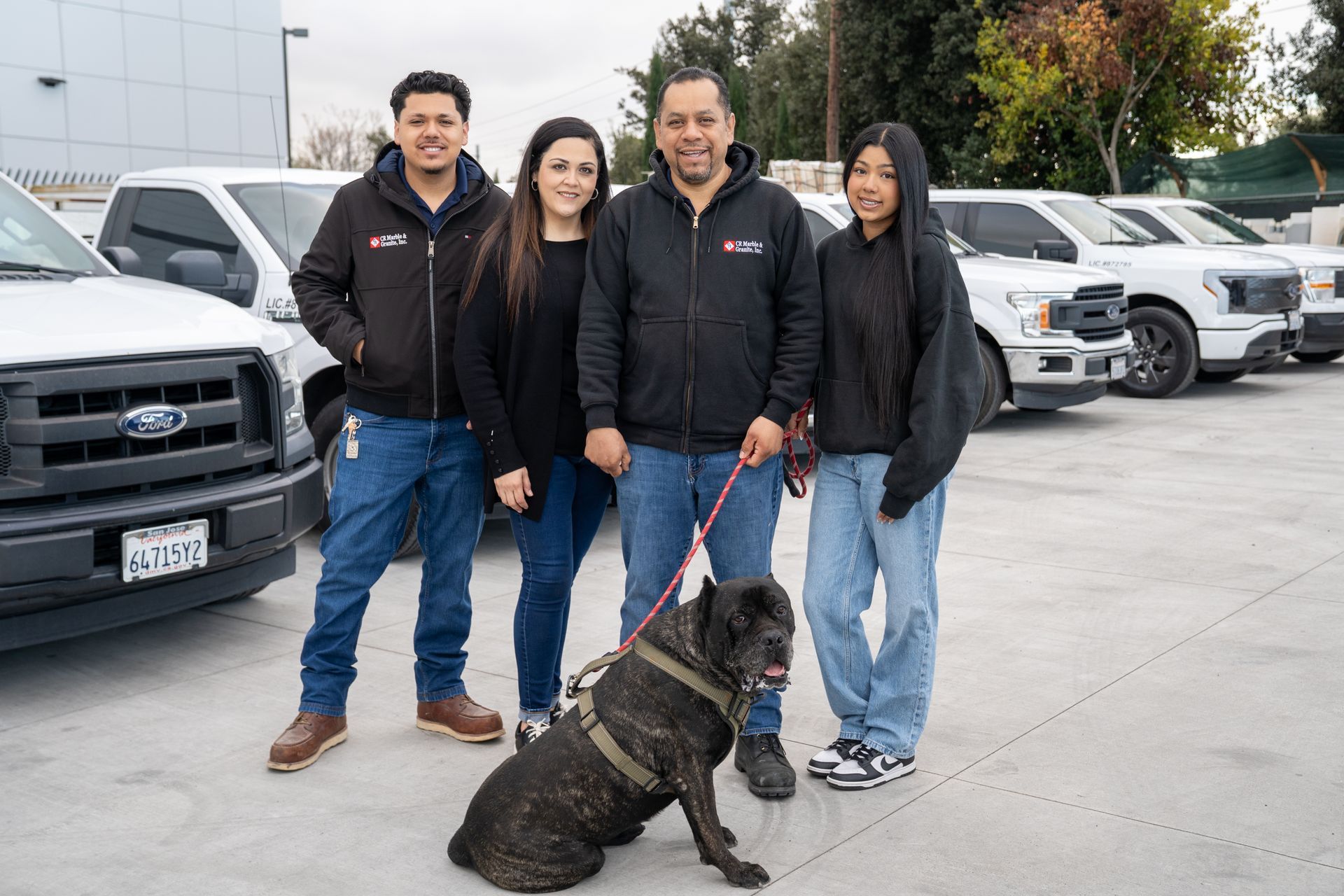 Family and dog pose in front of white trucks. They wear matching jackets and jeans.